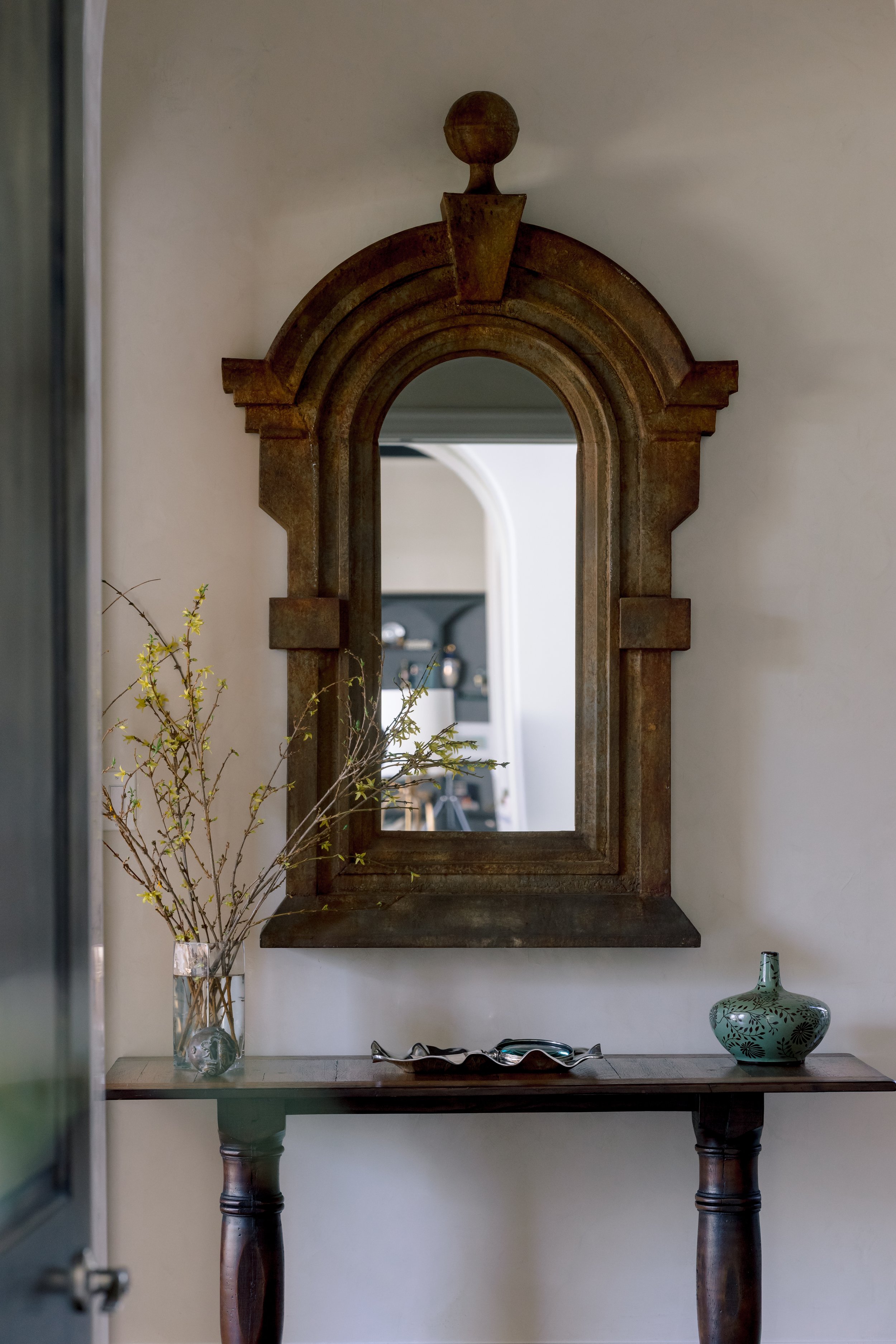 A wooden table with a decorative vase containing branches and a green vase, a mirror with a wooden frame hanging above, and a mirror reflecting part of a room, against a plain wall.