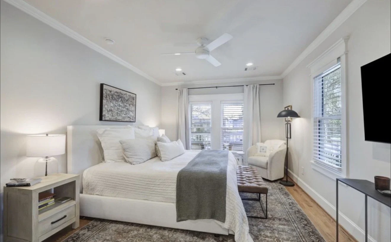 A bedroom with a white bed, gray blanket, bedside lamp, armchair, flat-screen TV, and windows with white curtains