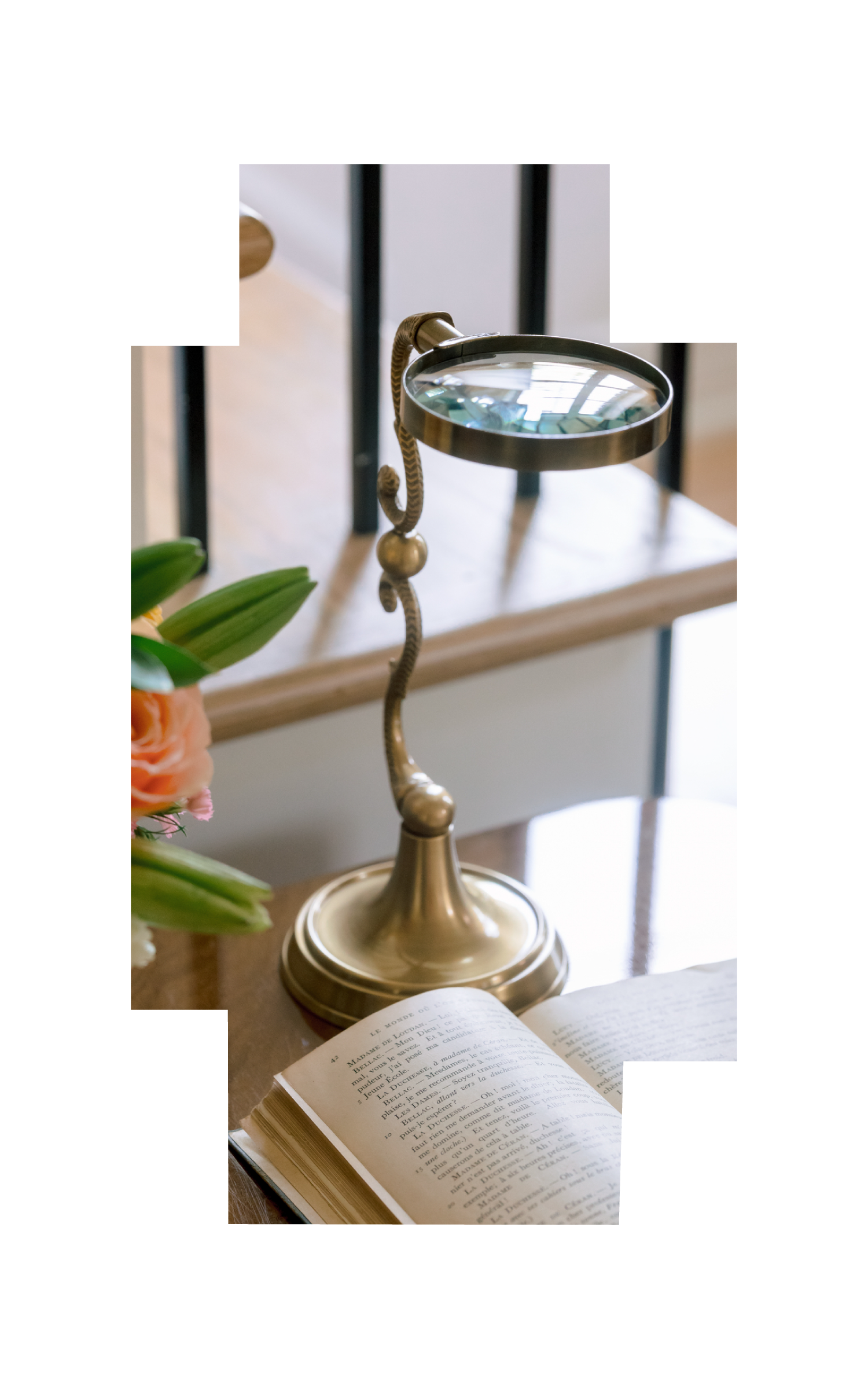 A brass magnifying glass with a curved handle is placed beside an open book on a wooden table. There is a flower arrangement with peach and pink flowers and green leaves to the left of the book. A white wall and a black metal railing are in the backg