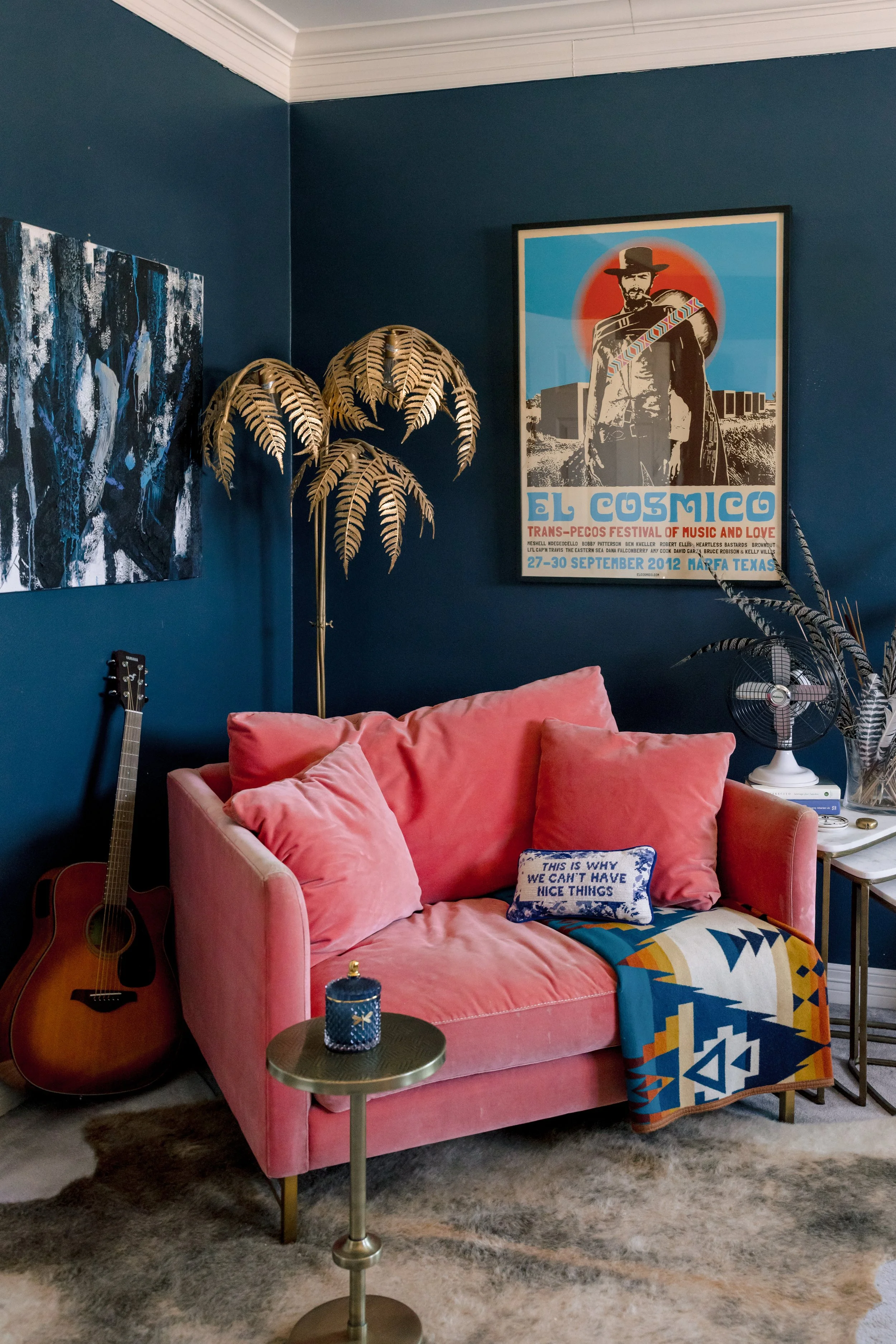 A cozy living room corner featuring a pink velvet couch with pillows and a patterned blanket, a guitar leaning against the wall, a small side table with a decorative item, a fan on a side table, artwork on the deep blue wall, a large plant with gold 