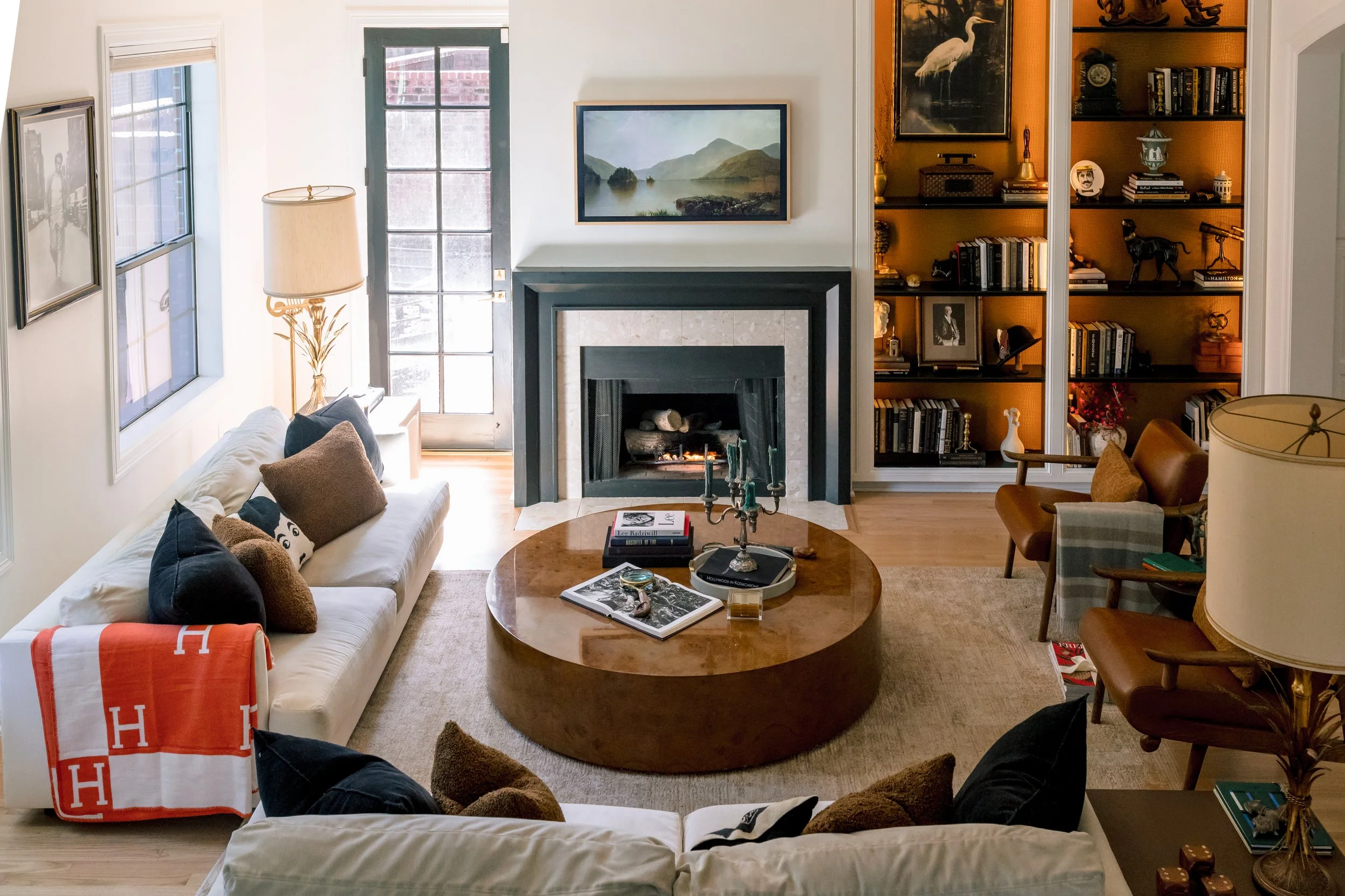 A living room with a white sofa on the left decorated with various pillows and a blanket. In the center, there's a round wooden coffee table with books, a candle holder, and a candelabrum. Behind the table, there's a fireplace with a framed landscape