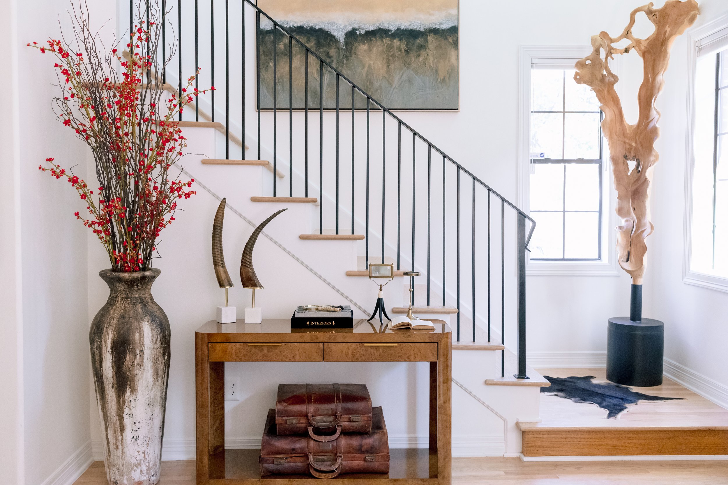 Interior entryway featuring a large vase with red berries and twigs, a wooden console table with books and decorative horns, and a tall, abstract wooden sculpture next to a window.