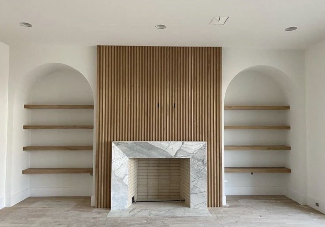 Living room with a fireplace surrounded by marble, flanked by oak wood shelves on each side, and a vertical oak wood slat wall above the fireplace.