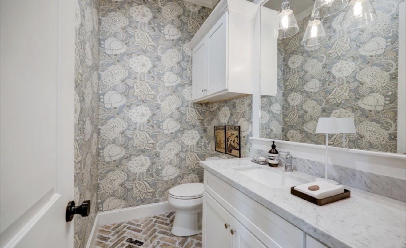 A small bathroom with floral wallpaper, a white marble vanity with a bottle of soap, a lamp, and framed pictures. There is a white toilet and a large mirror above the vanity. The flooring is made of brick-style tiles.