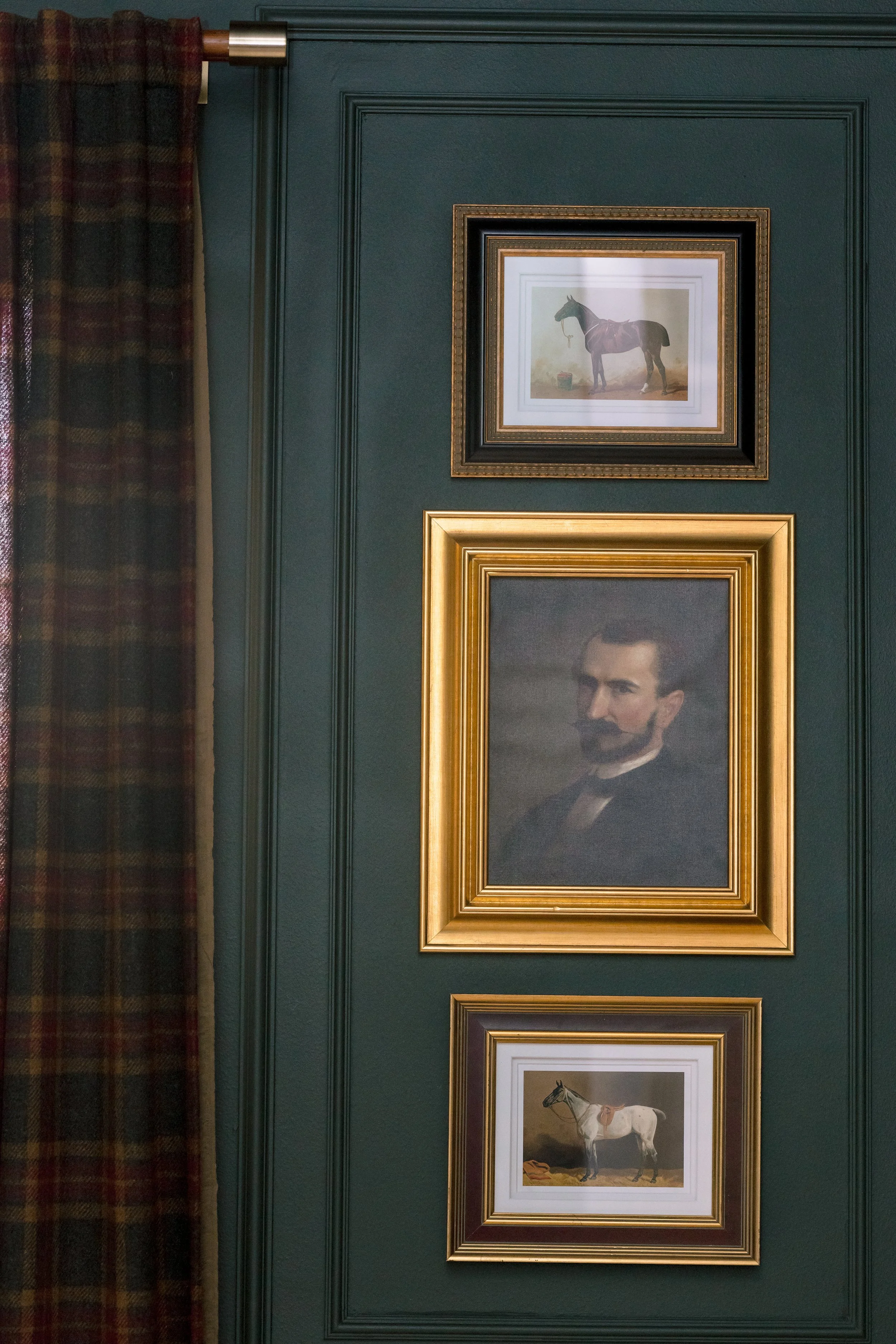 A green wall with a framed painting of a horse, a framed portrait of a man, and a framed painting of a horse, with a plaid curtain on the left side.