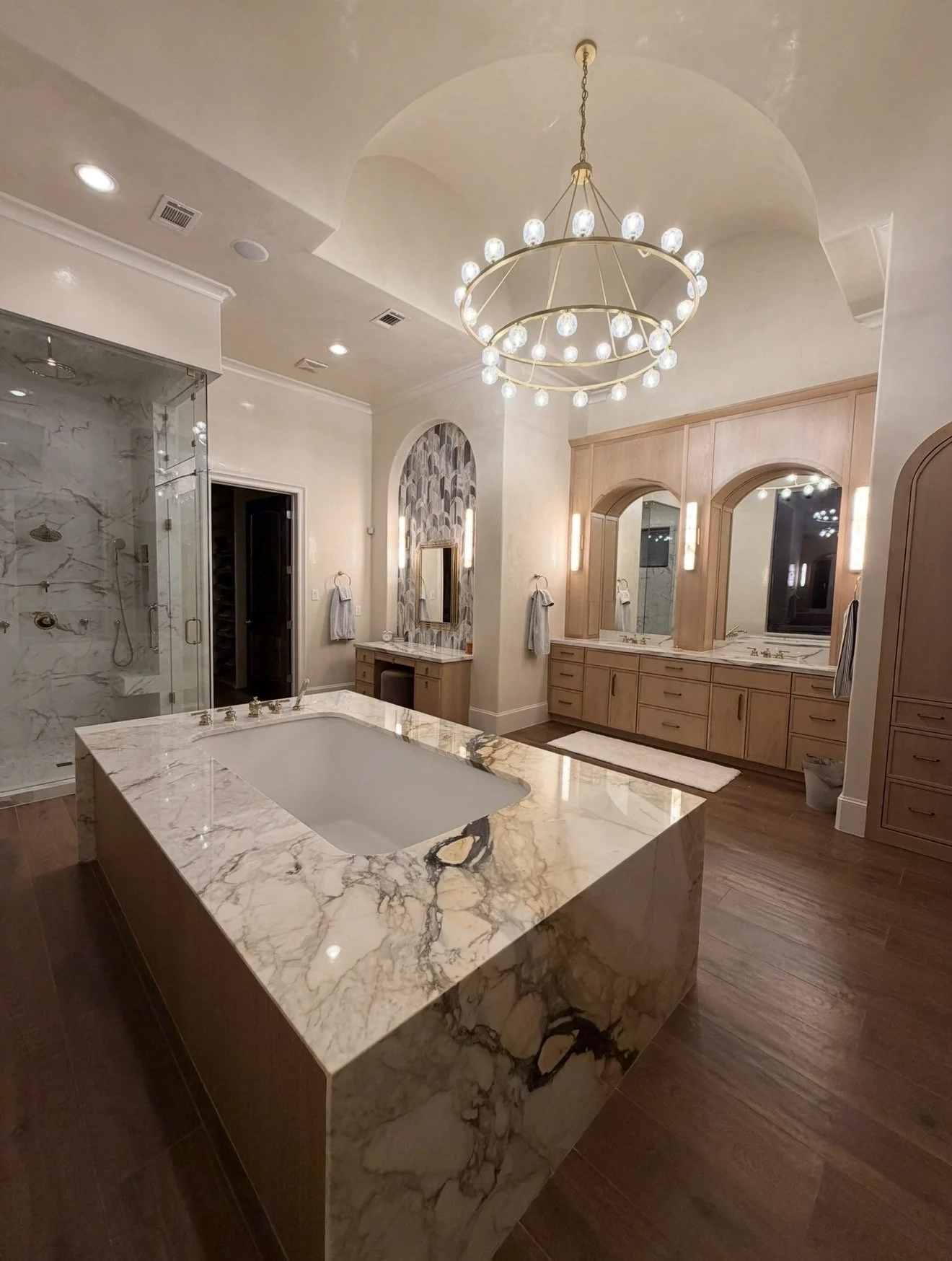 Elegant bathroom featuring a large marble-topped bathtub, double vanities with mirrors, hardwood floors, a walk-in shower with marble walls, and a modern chandelier.