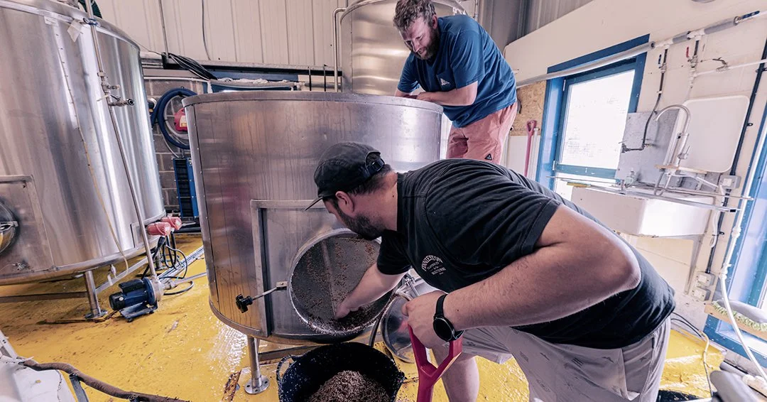 Two men working in a brewery, one pouring roasted grains into a large stainless steel brewing vessel, the other leaning inside the vessel, both engaged in brewing beer.