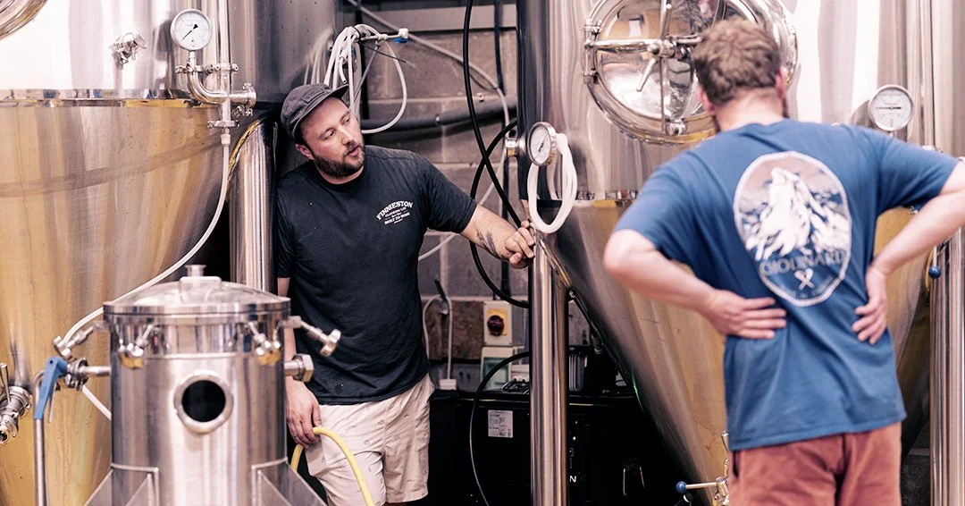 Two men working in a brewery with large stainless steel fermentation tanks.