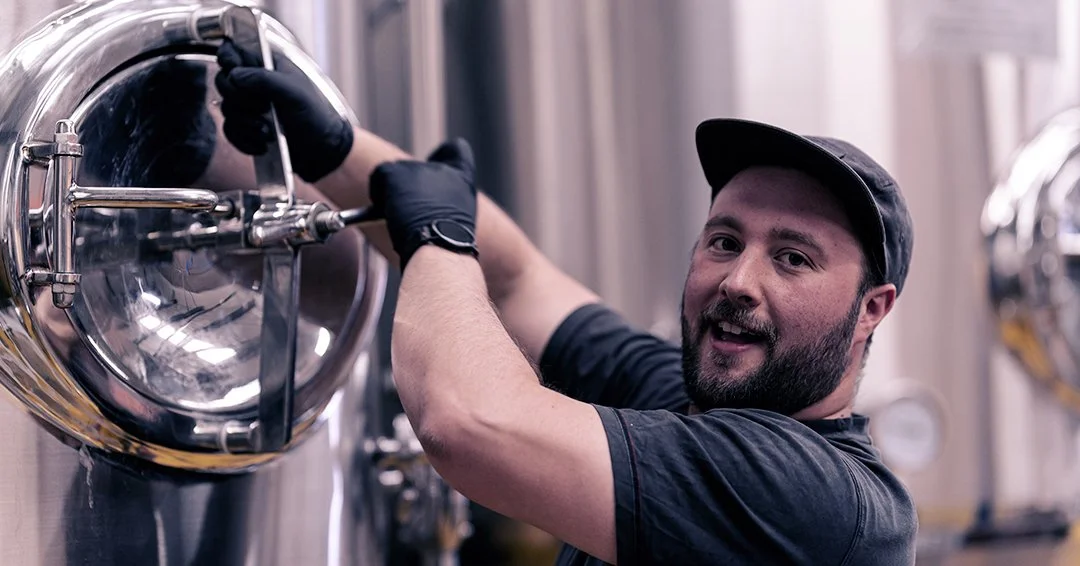 A man wearing a black cap and black gloves is inspecting or working on a large, shiny metal fermentation tank in a brewery or industrial setting, smiling at the camera.