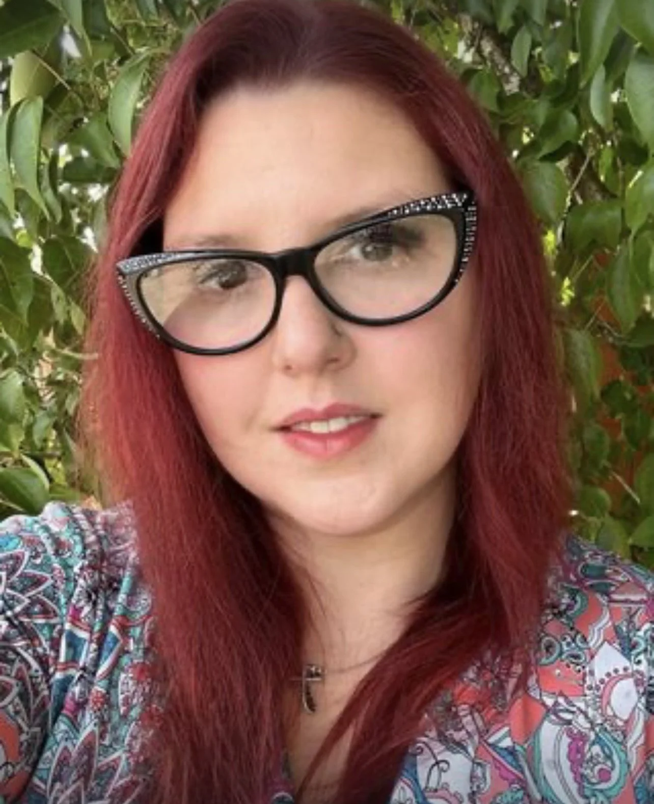 A woman with red hair, wearing black-rimmed glasses with rhinestones, smiling outdoors in front of green foliage.