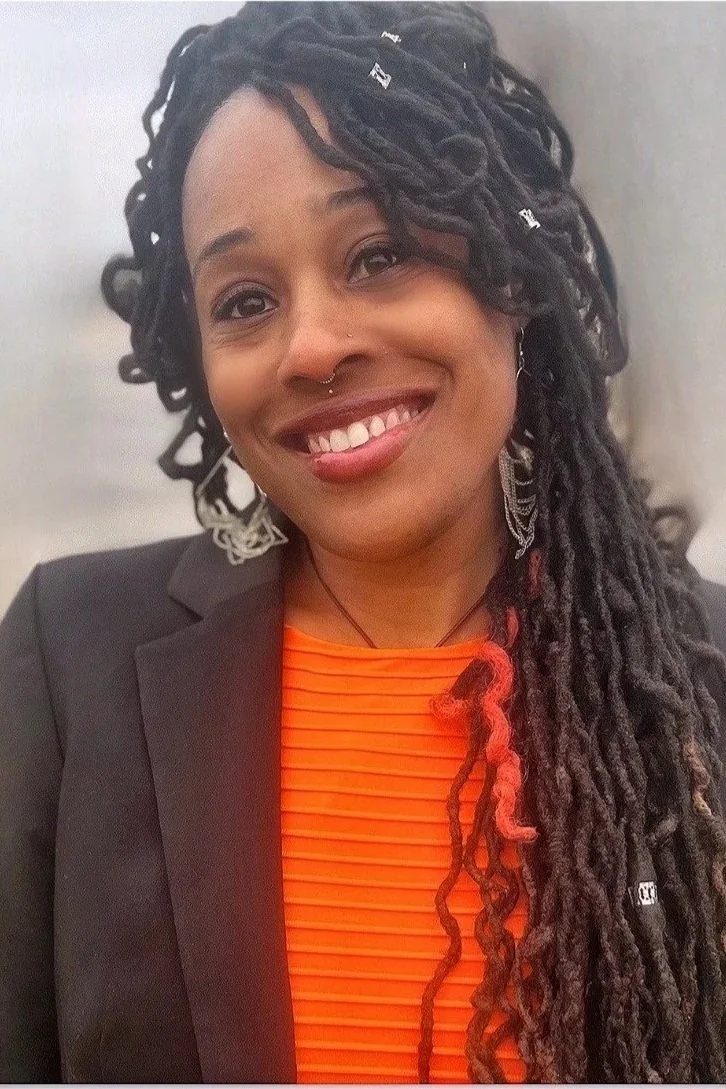 A woman with long dreadlocks, some tied with small silver accessories, smiling, wearing a black blazer over an orange top, with earrings and a nose ring.