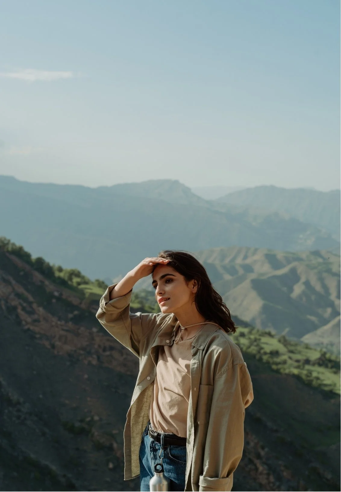 A woman standing outdoors with mountains and greenery in the background, shielding her eyes from the sun with her hand.