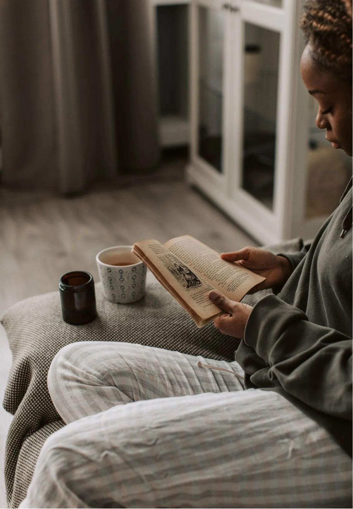 A person sitting on a couch reading a book with a cup of tea and a small dark bottle on a side table in a cozy living room.
