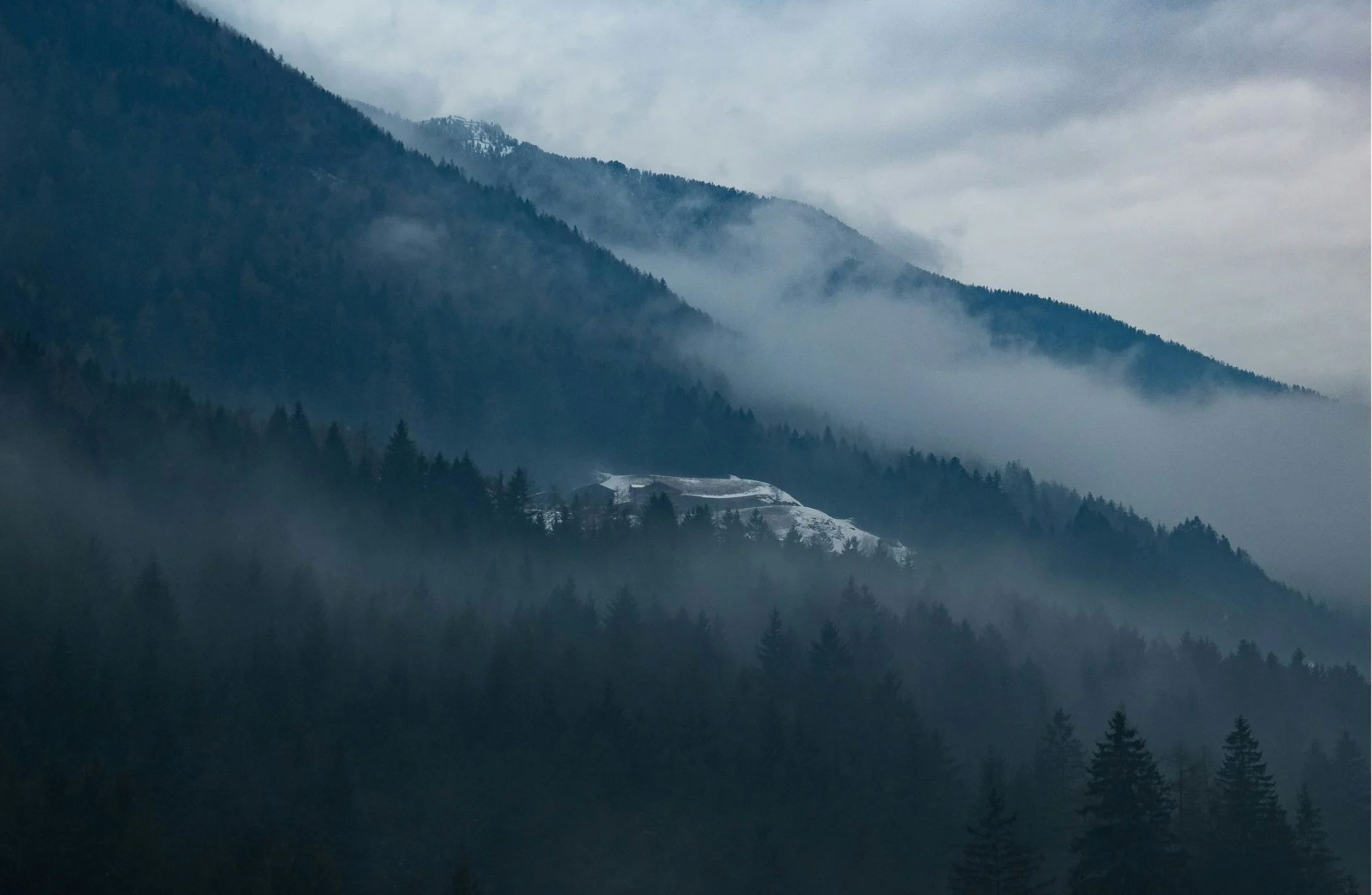 Mountain landscape with forested slopes and misty clouds, snow-covered building on a hillside.