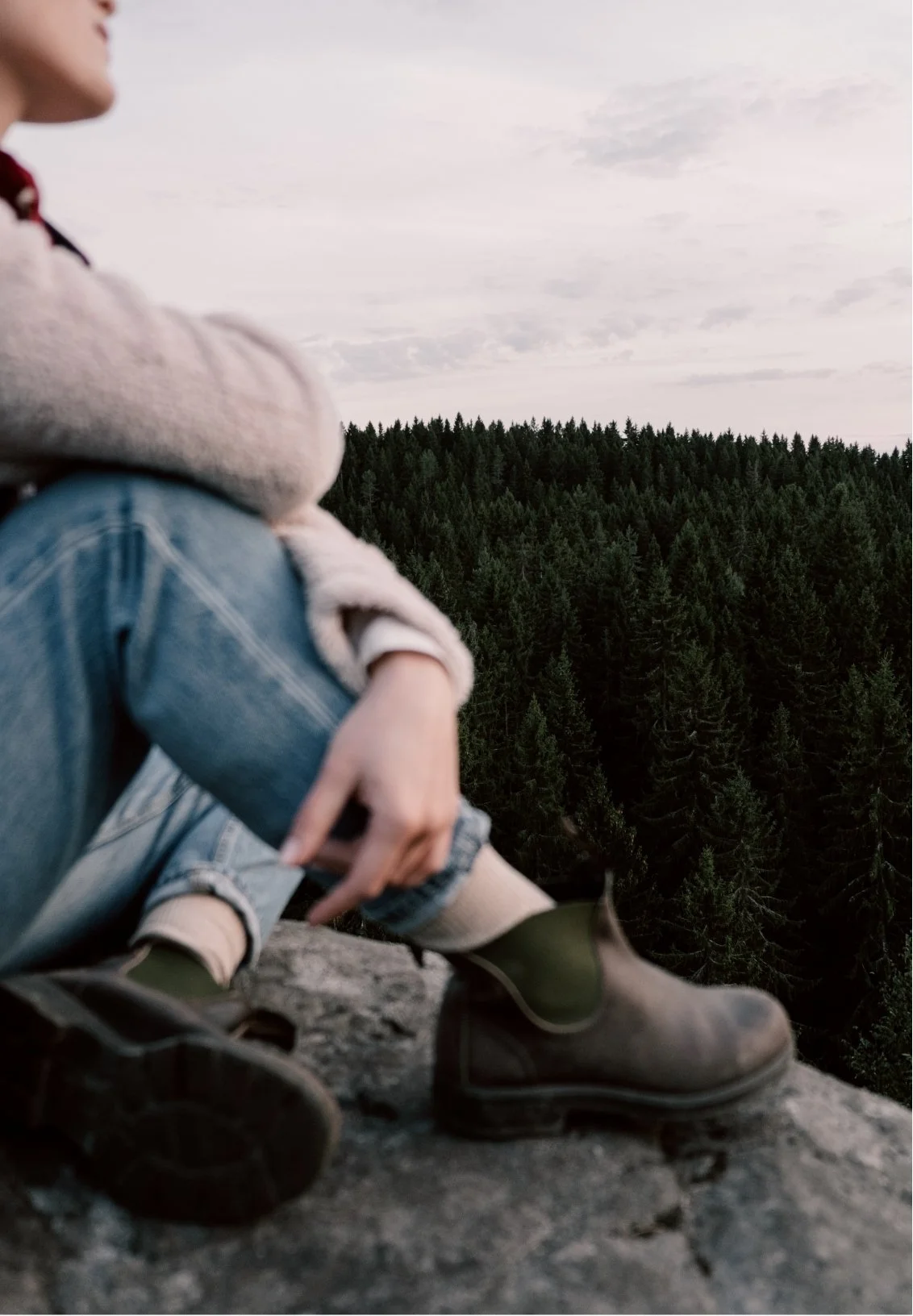 Person sitting on a rocky ledge overlooking a dense forest with cloudy sky.
