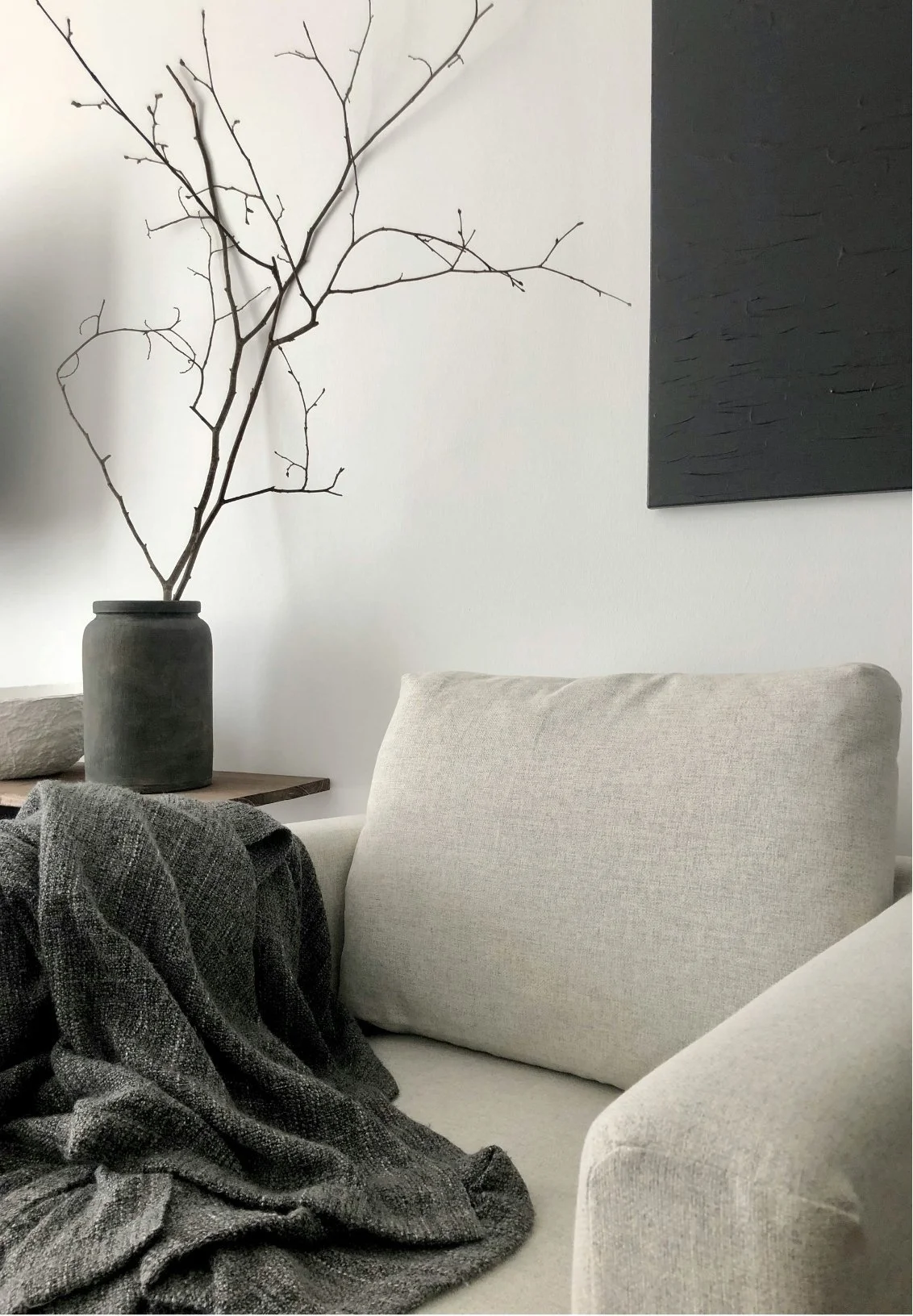 Interior living room scene with a beige armchair, a dark blanket draped over it, a wooden side table, a large black vase with bare branches, and a textured dark wall art piece on a white wall.