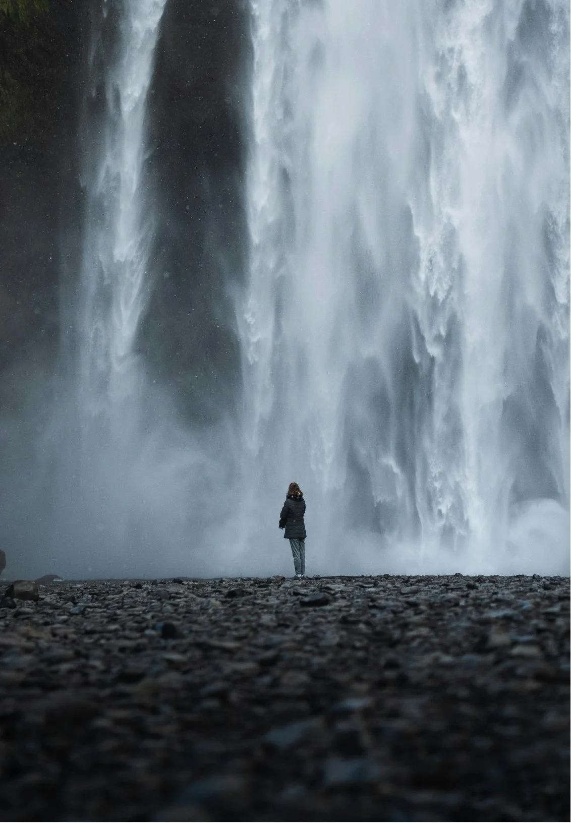 A person standing on a rocky ground in front of a large waterfall with tall, cascading water.