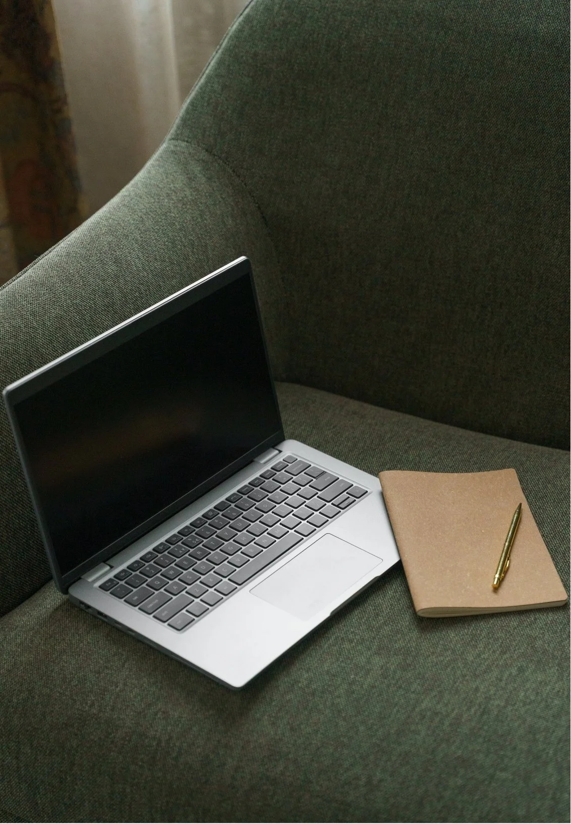 Open silver laptop, closed notebook, and gold pen on a green fabric couch.