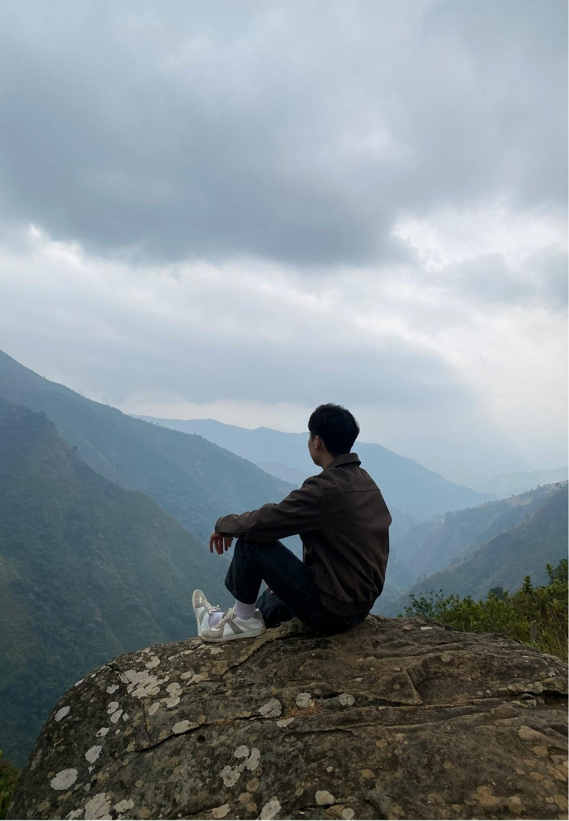 A person sitting on a large rock, looking out over a misty mountain landscape under cloudy skies.