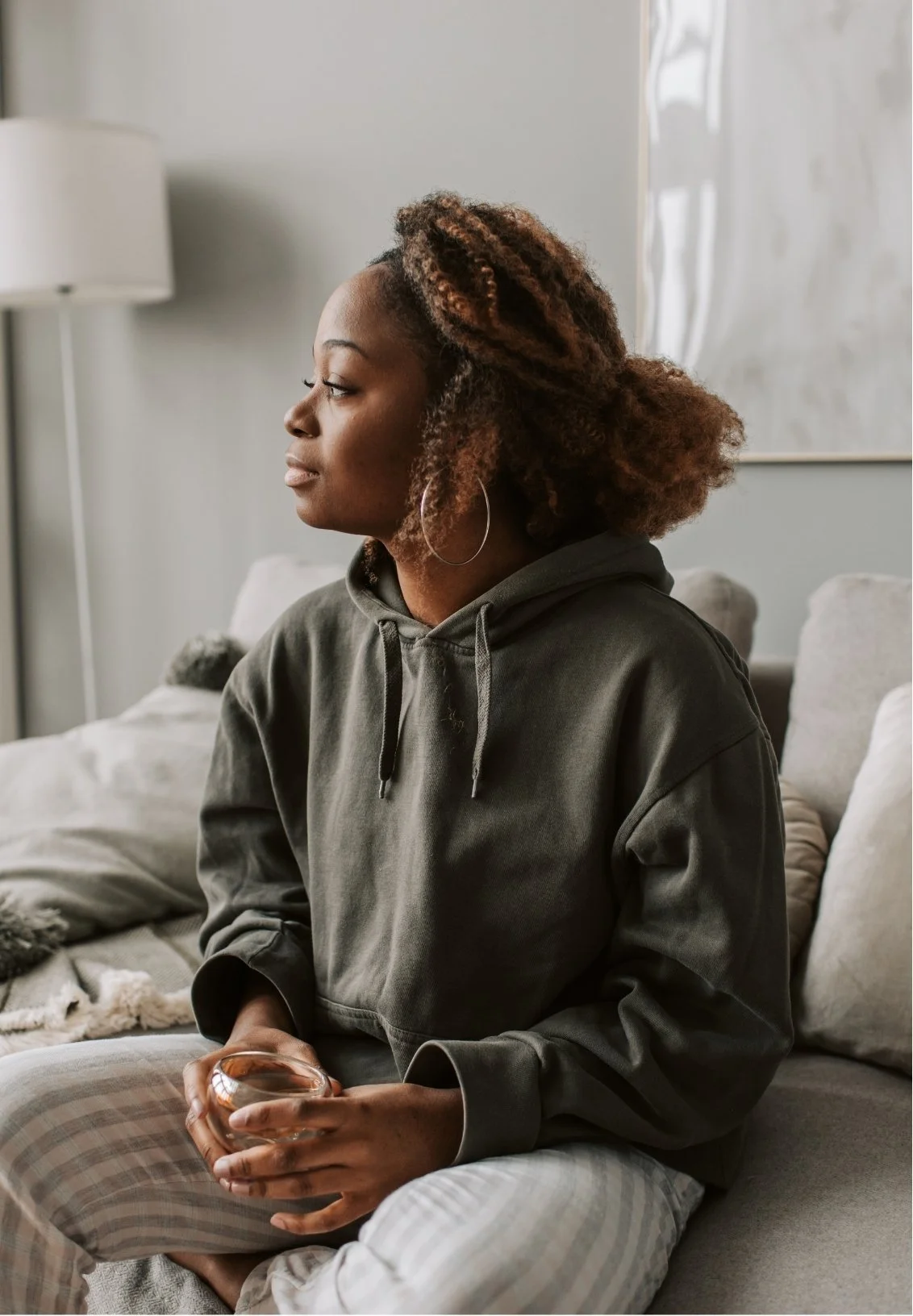 A woman with curly brown hair wearing a gray hoodie and plaid pajama pants sitting on a sofa, looking pensively to her left, holding a glass of water in her hands at home.