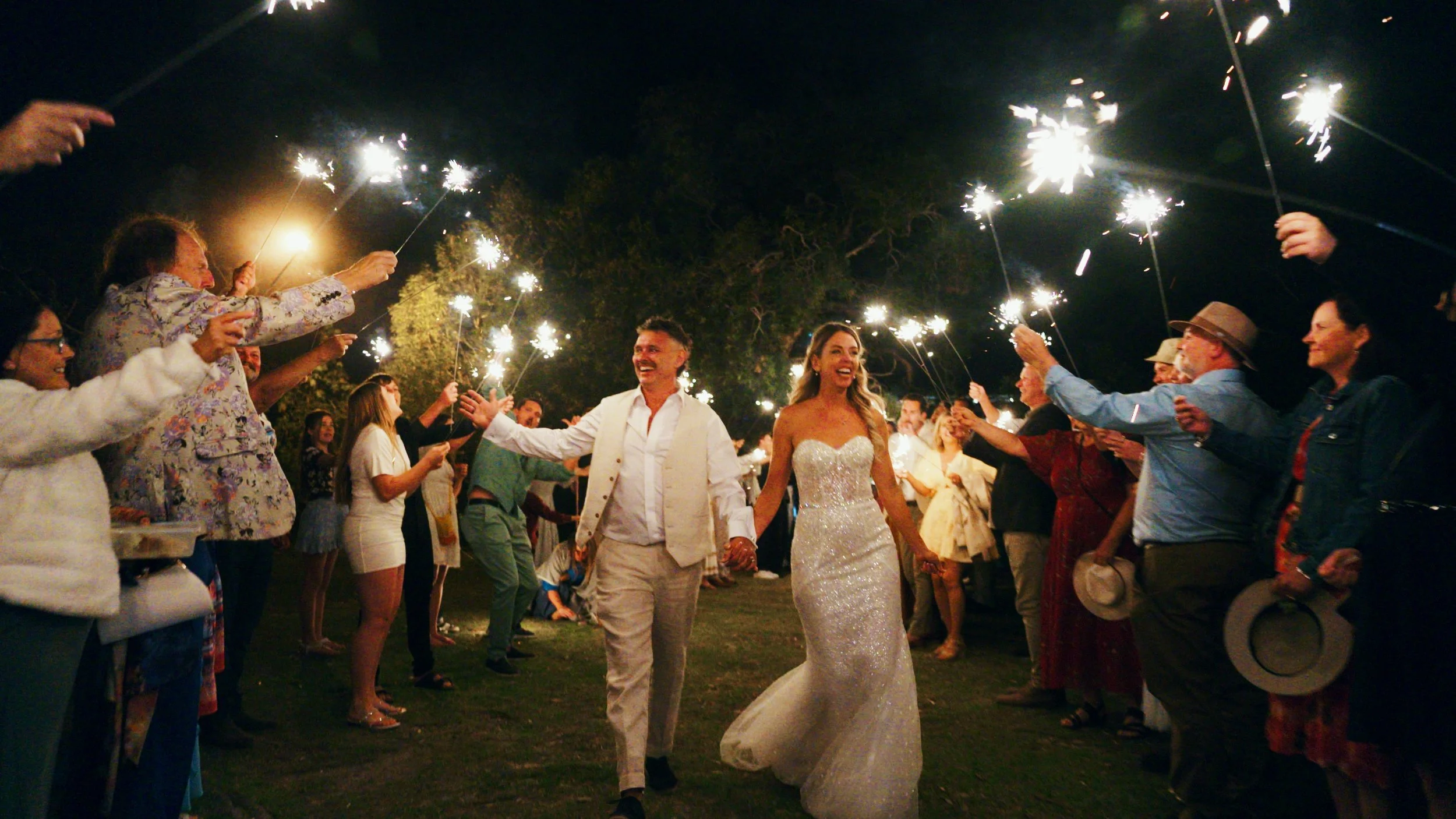 Wedding celebration at night with the bride and groom holding hands and walking through guests holding sparklers.