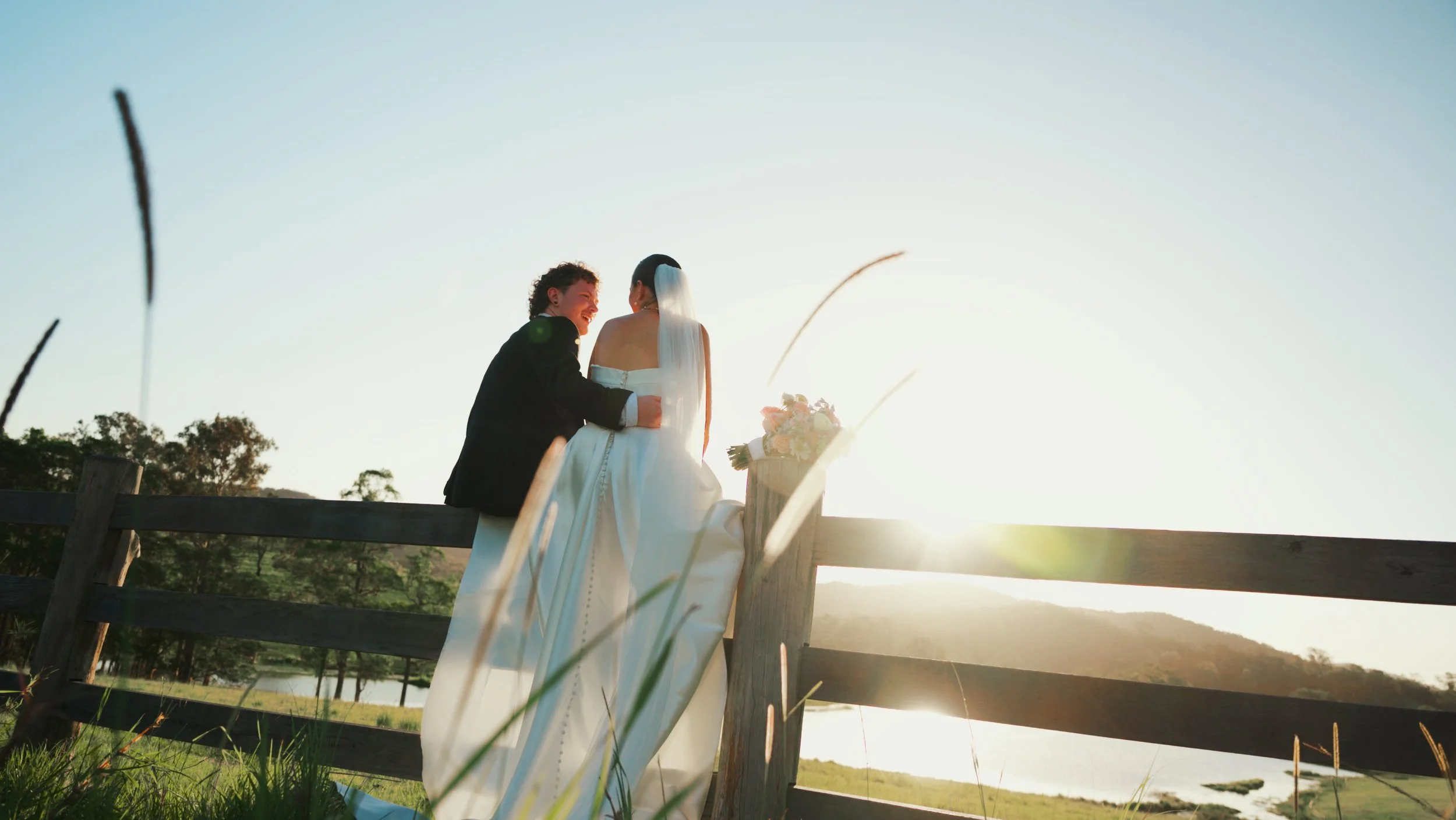 A newlywed couple sitting on a wooden fence in a scenic outdoor setting during sunset, with a bouquet of flowers nearby.