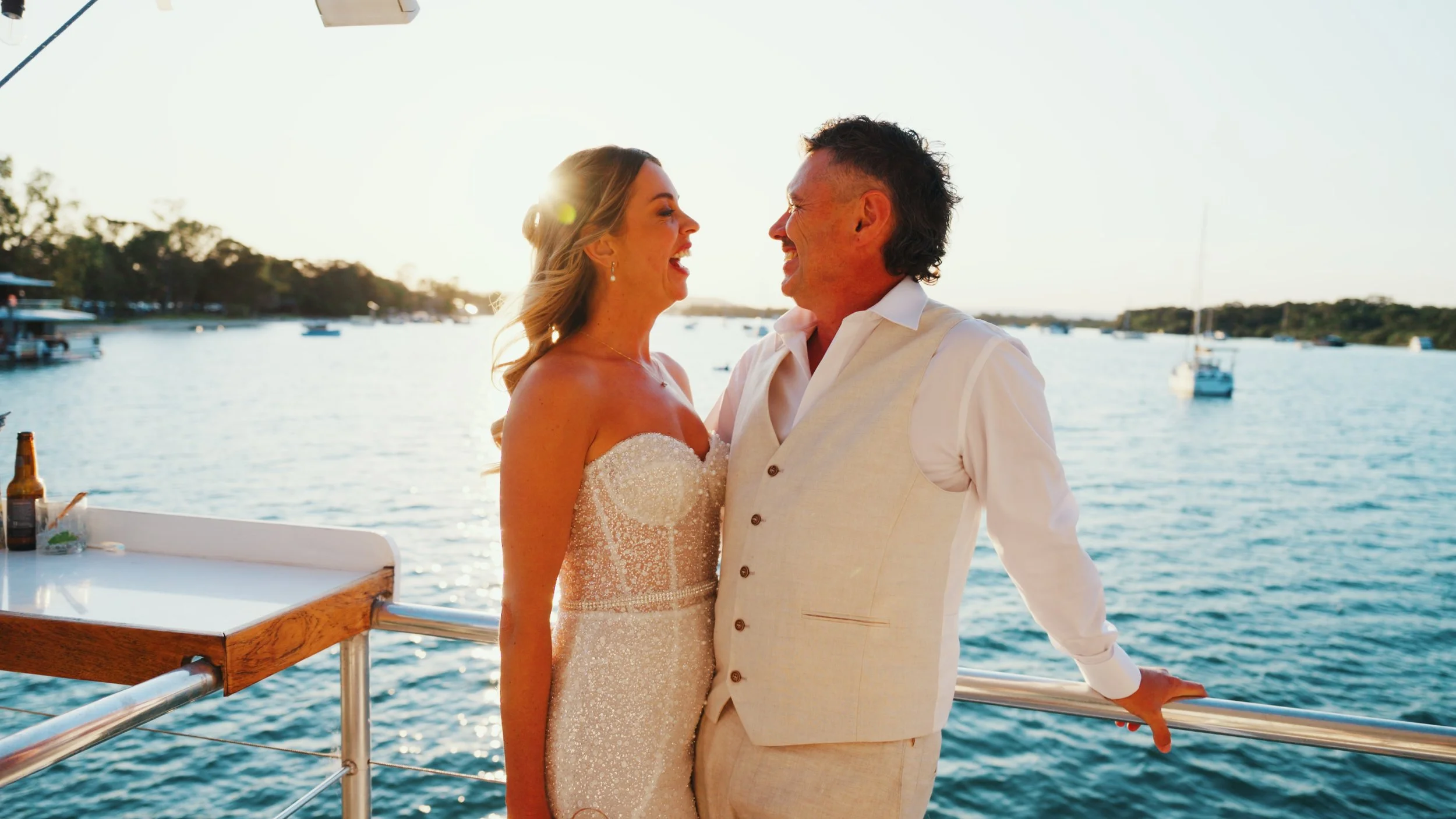 A couple dressed in wedding attire standing on a boat with a waterway and boats in the background, smiling and looking at each other at sunset.