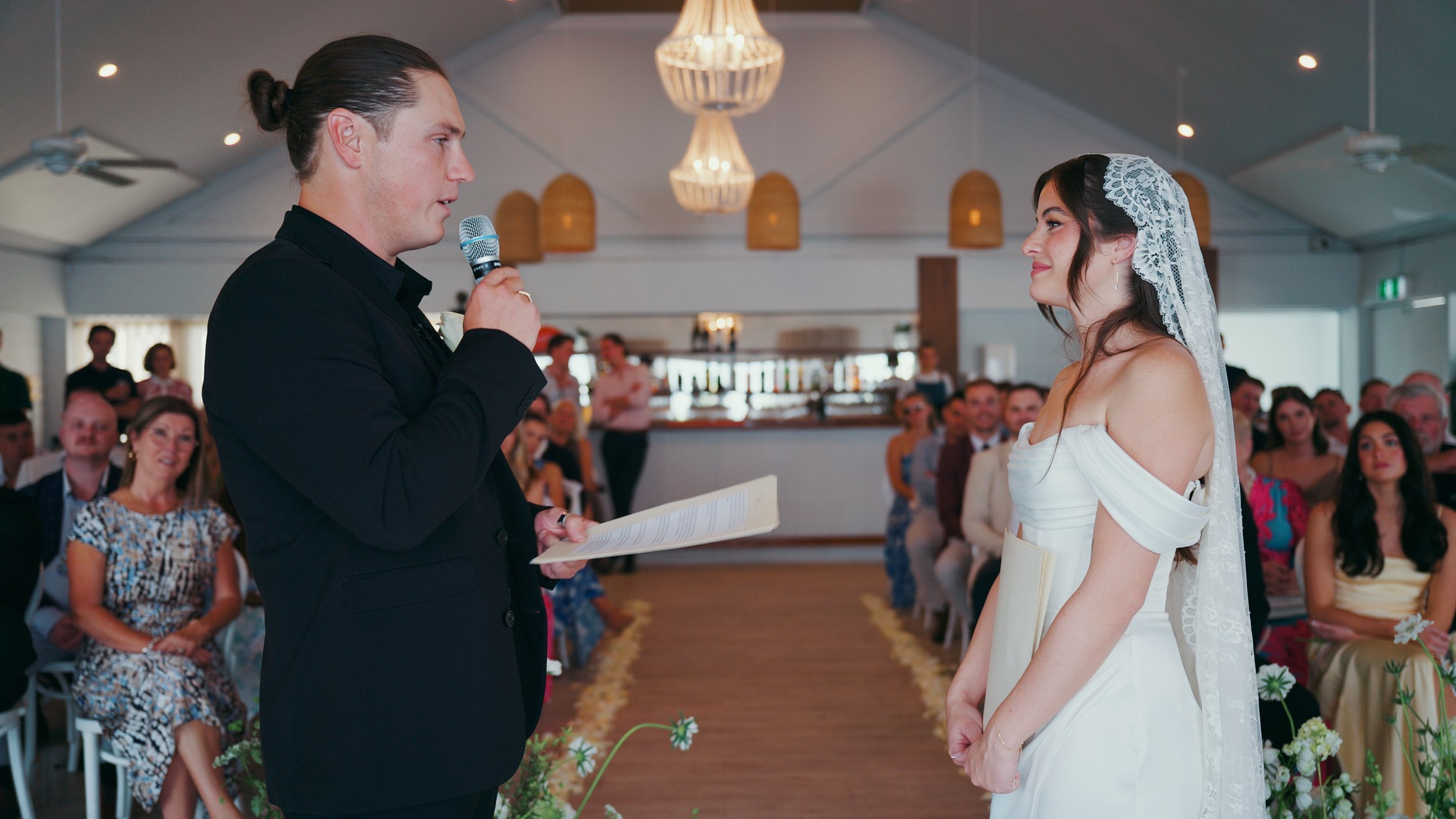 A groom in a black suit holding a microphone and reading vows to a bride in a white wedding dress with lace veil, during a wedding ceremony in a decorated indoor venue with seated guests watching.