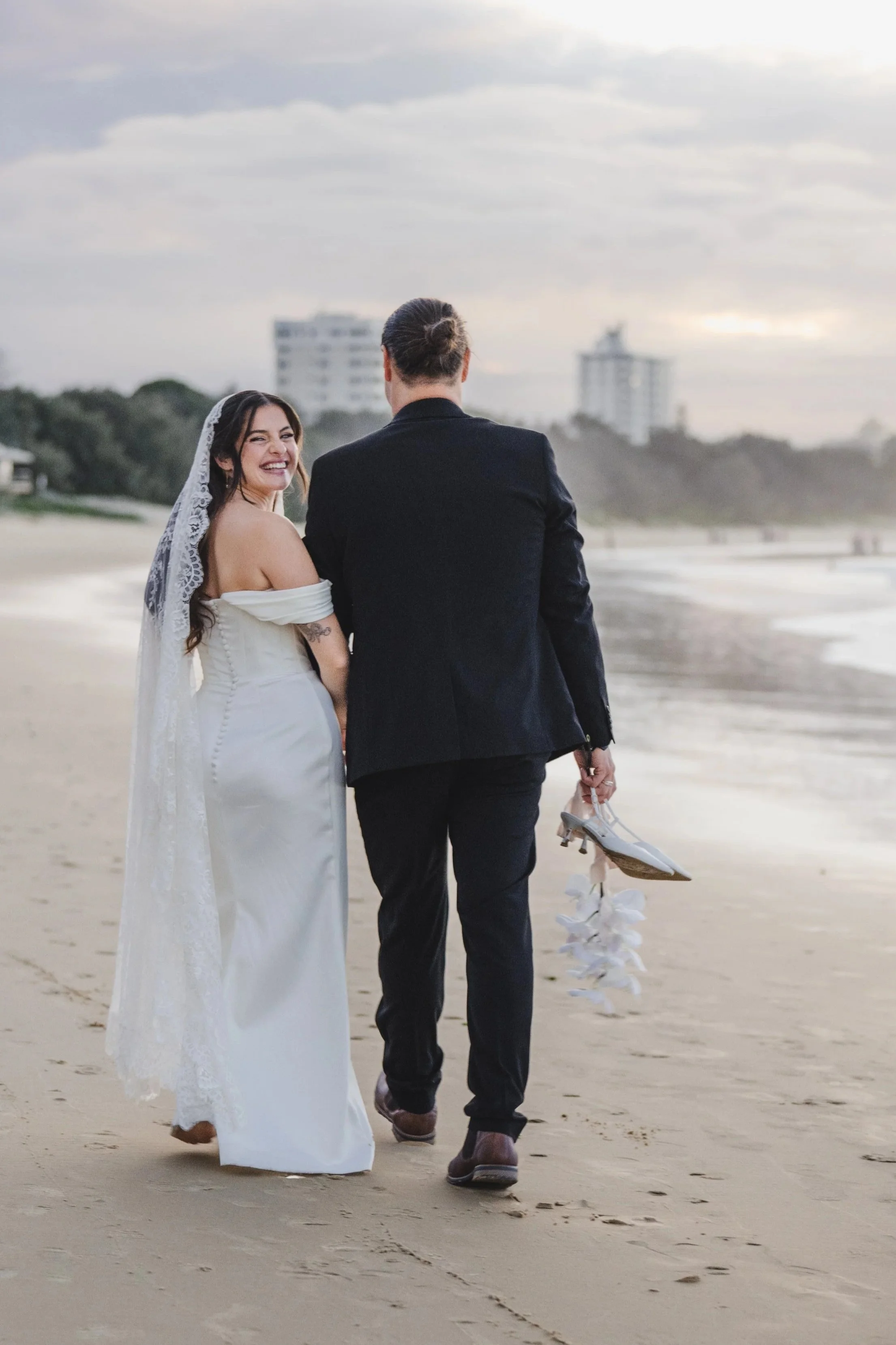 A bride and groom walking on the beach during sunset, with the bride smiling and holding her wedding shoes in her hand.