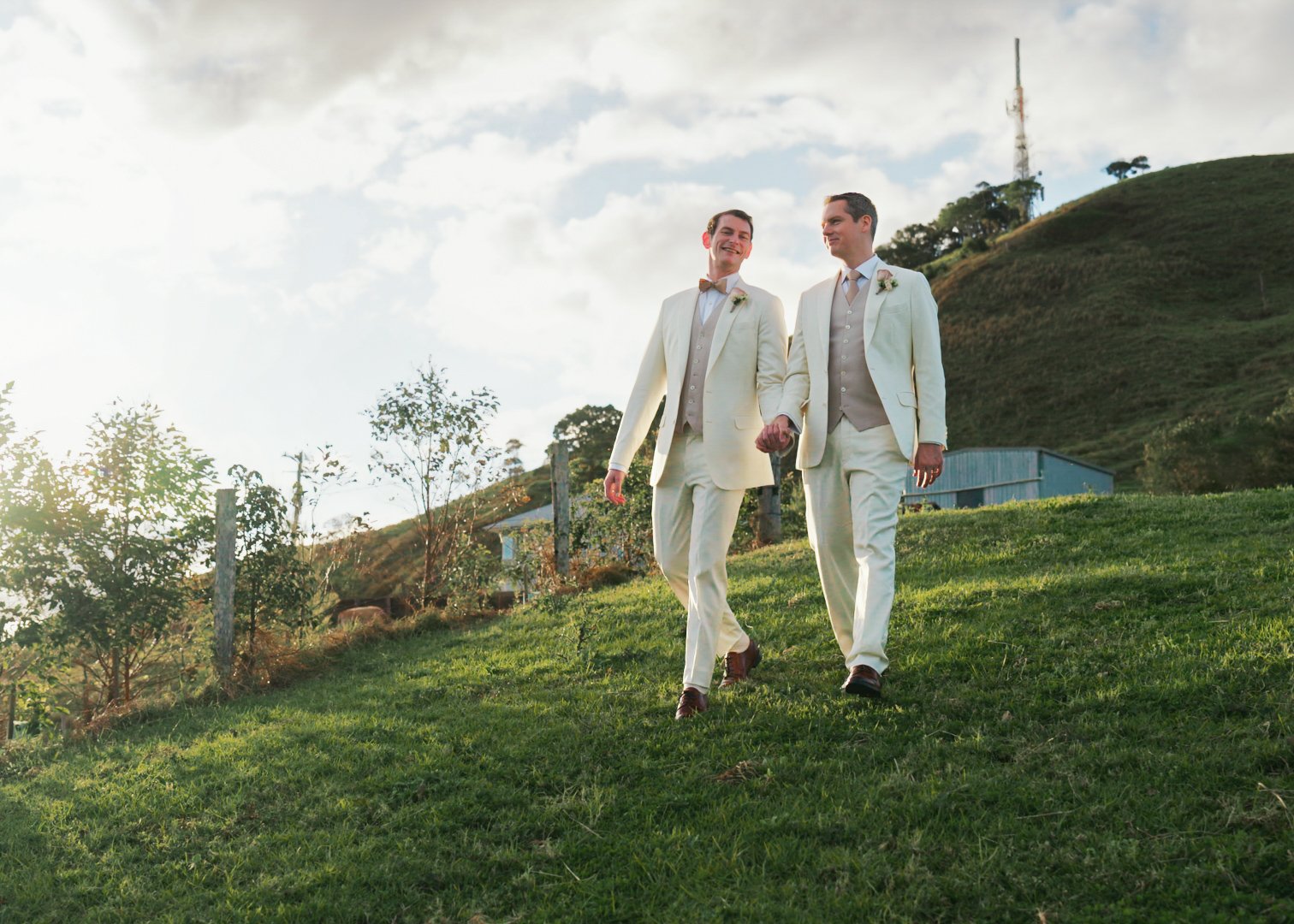 Two men in beige suits holding hands and walking on a grassy hill in a rural area with trees, a fence, and a small building in the background, under a partly cloudy sky.