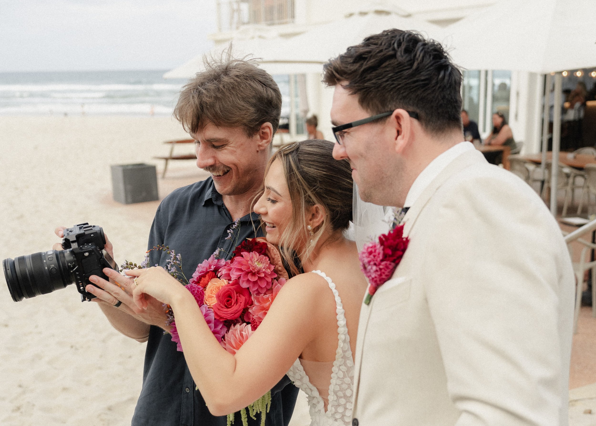 A wedding ceremony on the beach with a bride in a white dress holding a bouquet of red and pink flowers, standing next to a groom in a white suit with a pink boutonniere, while a man in a dark shirt shows them a photograph on his camera.