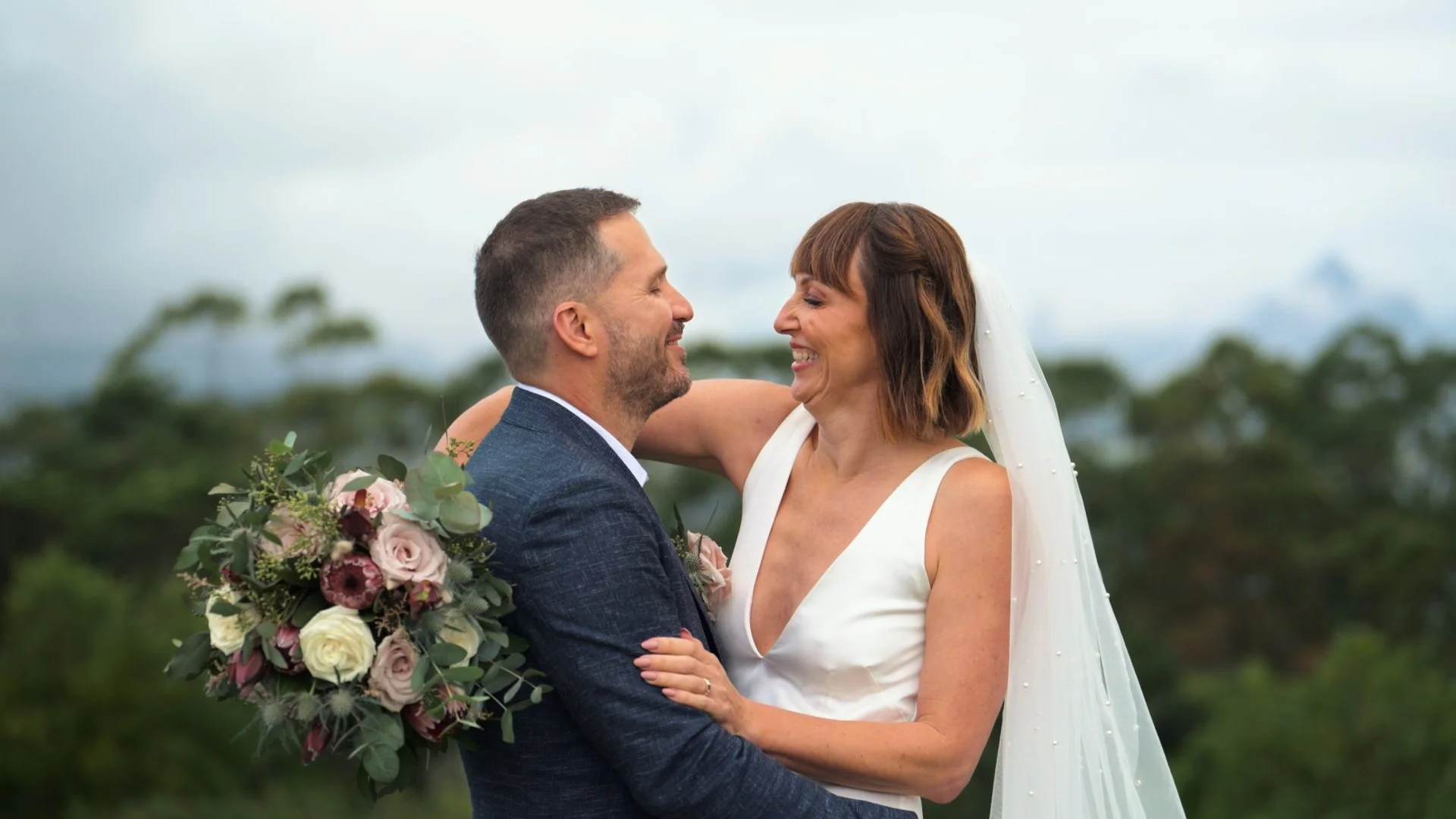 A bride and groom embrace outdoors with trees and cloudy sky in the background, smiling at each other, the bride holding a bouquet of pink, white, and purple flowers.