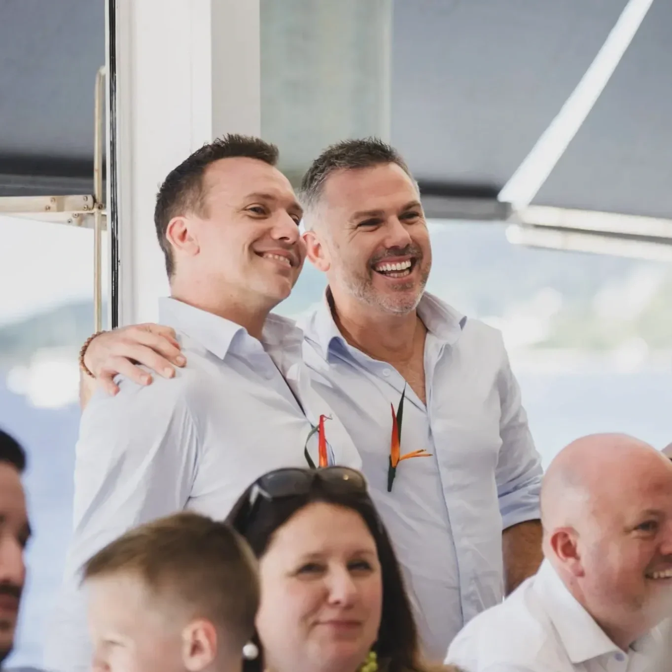 Two smiling men in white shirts at a social gathering, with glasses hanging from their shirts, standing close together, in a bright room with large windows.