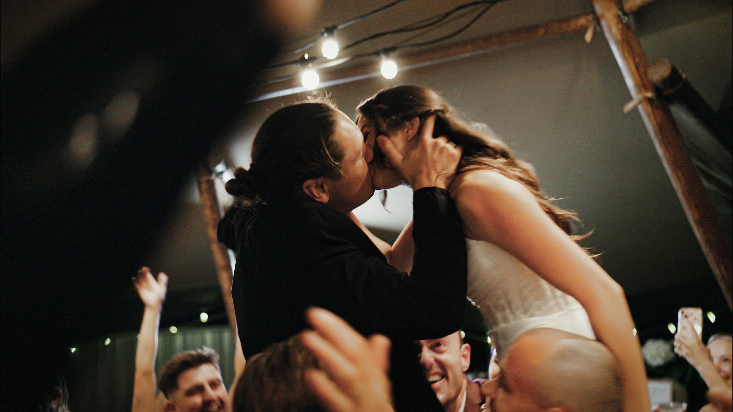 Couple kissing during a celebration, with onlookers smiling and raising their hands, in a warmly lit indoor space.