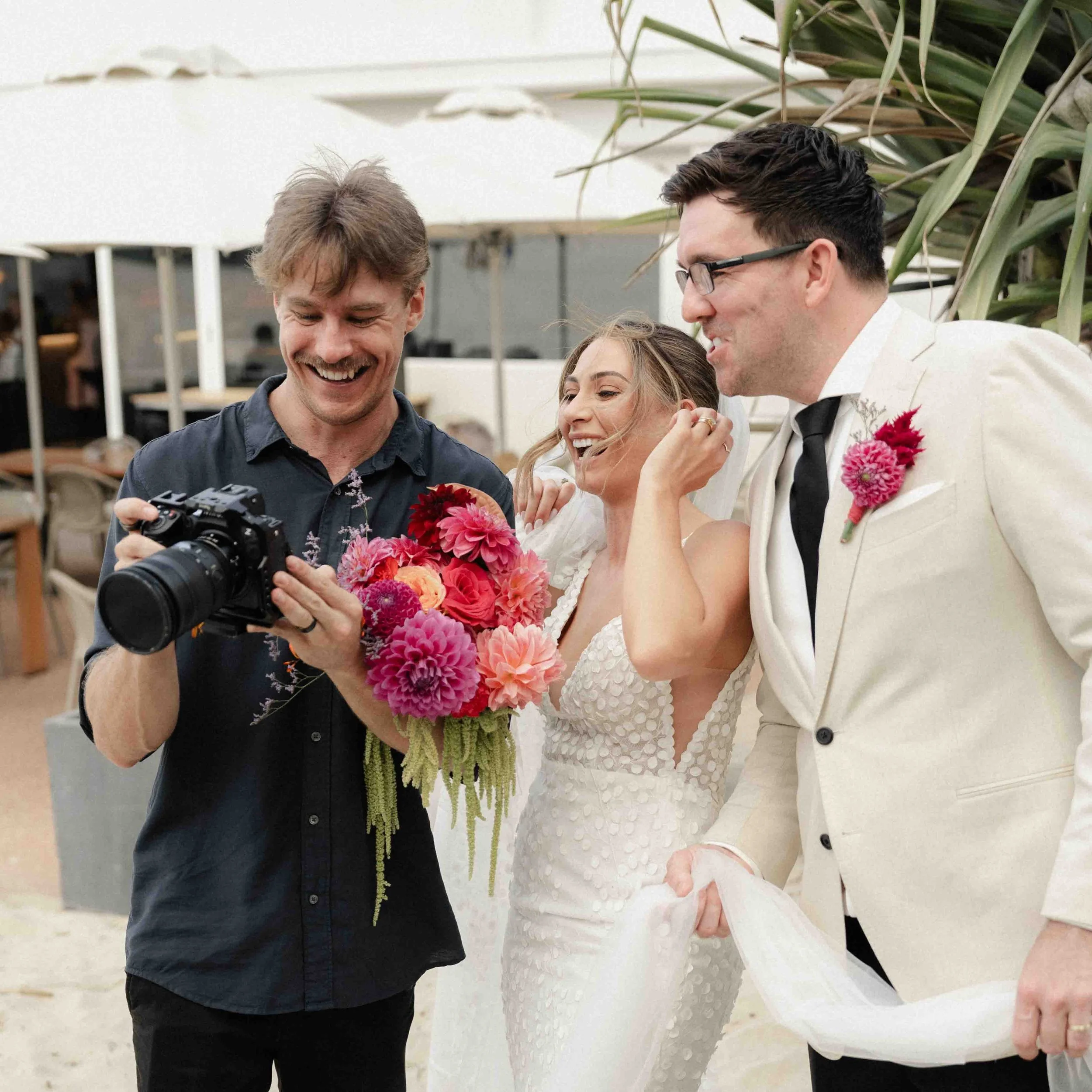 A bride in a white dress holding a colorful bouquet of pink and orange flowers, standing next to a groom in a white suit with a pink boutonniere, at a wedding reception. A man with a camera is showing something to the couple, with all three smiling and standing outdoors.