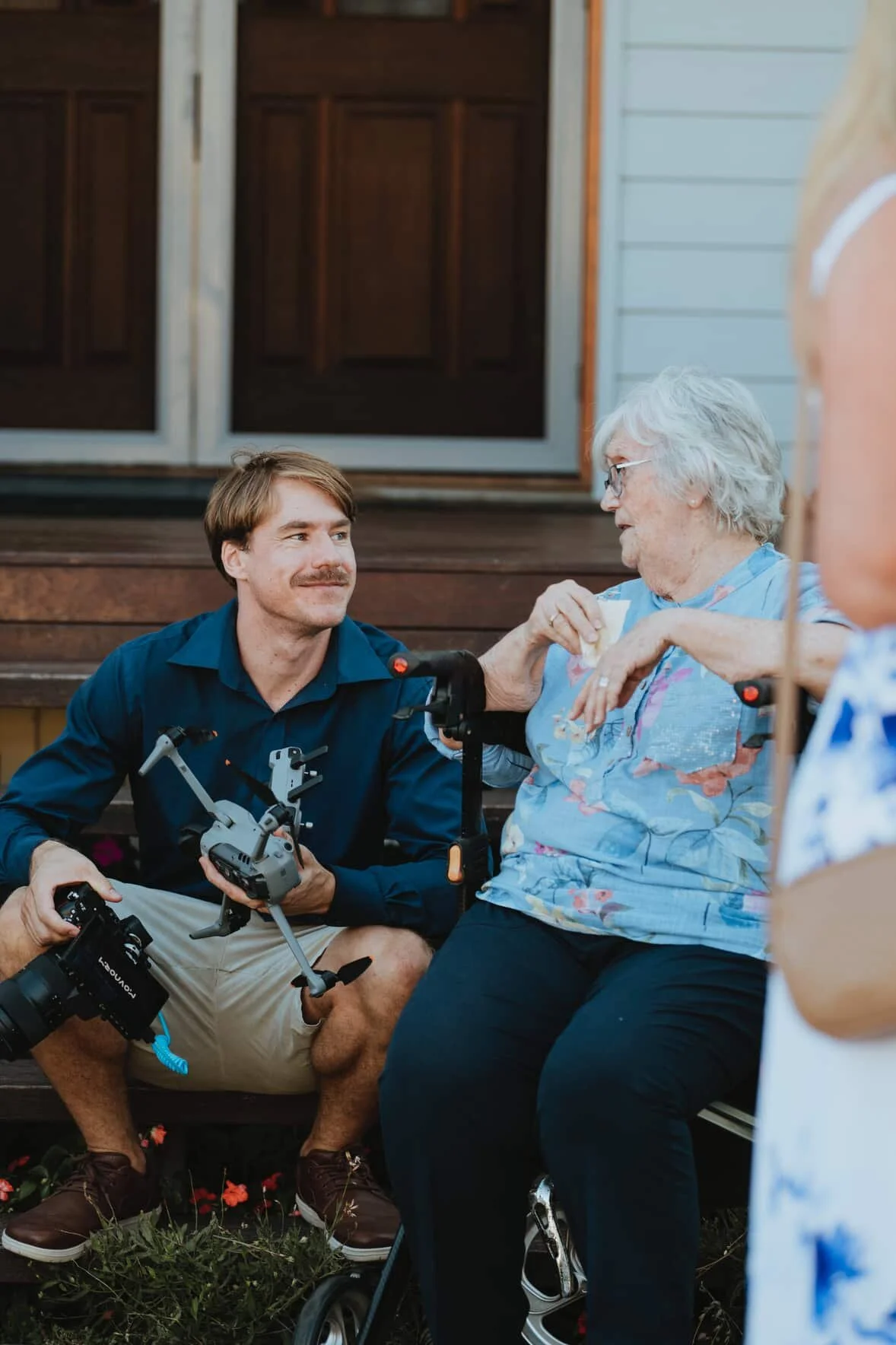 A young man with a drone and a camera sits next to an elderly woman in a wheelchair, sharing a moment on a front porch, with another person partially visible on the right.