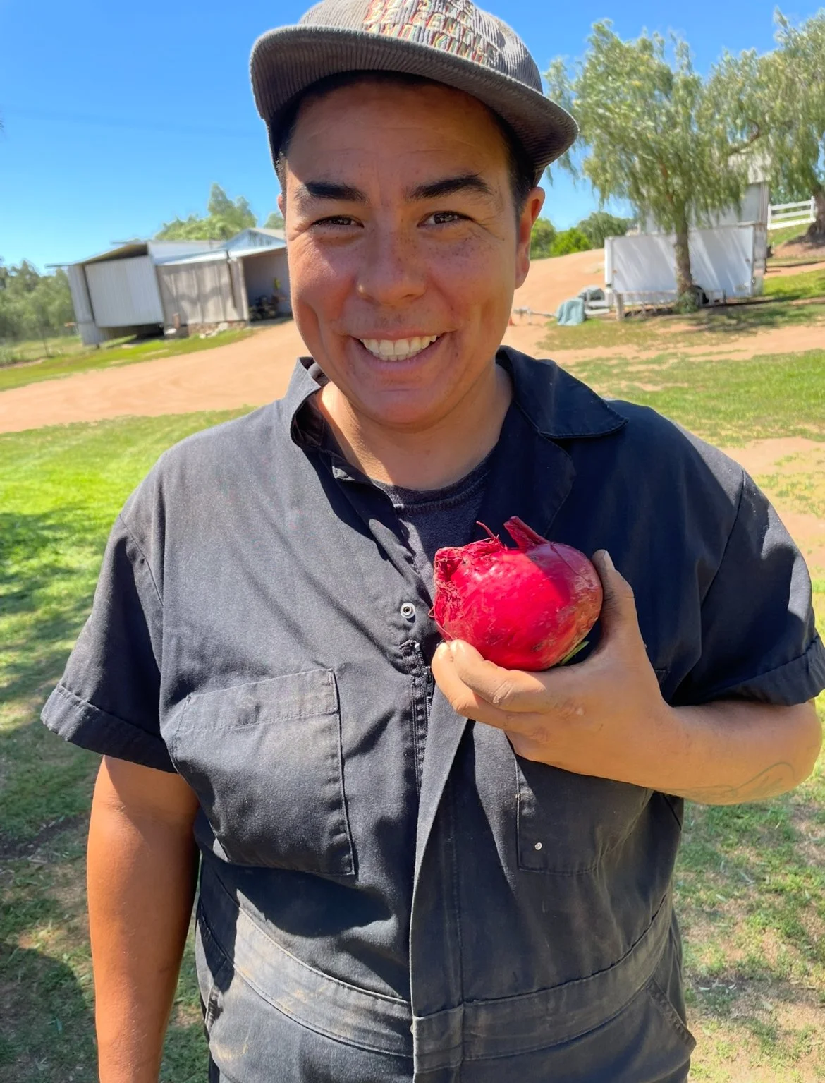 Ayzsha Smith, sole farmer of Kind Folk Farm based in San Diego is smiling outdoors holding a large red beet in their hand.