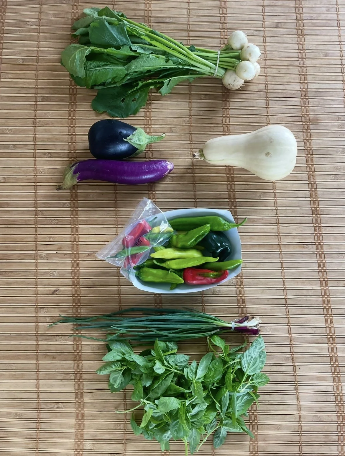 Assorted fresh vegetables and herbs from Kind Folk Farm's CSA on a bamboo mat, including green onions, turnips, eggplant, garlic, chili peppers, basil, and possibly some cherry tomatoes.