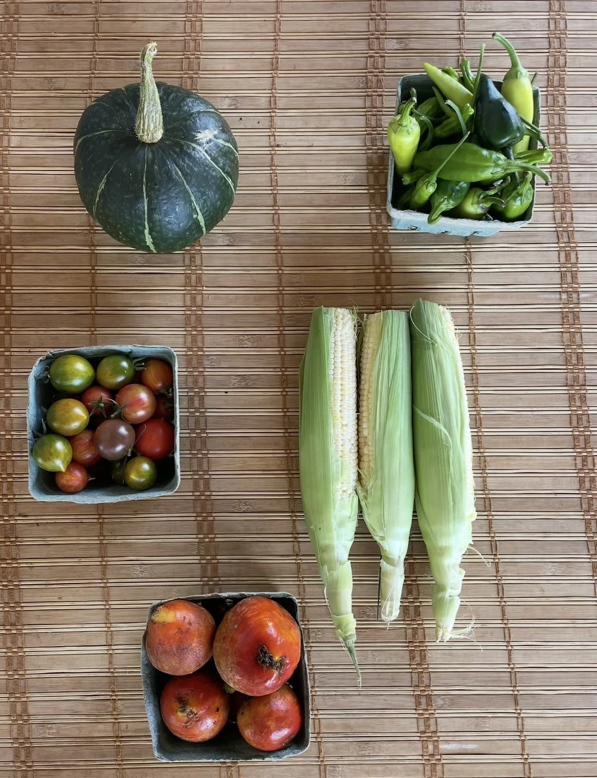 Arrangement of fresh vegetables from Kind Folk Farm's CSA on a bamboo mat, including a green pumpkin, green chili peppers, heirloom cherry tomatoes, three ears of corn, and pomegranates.