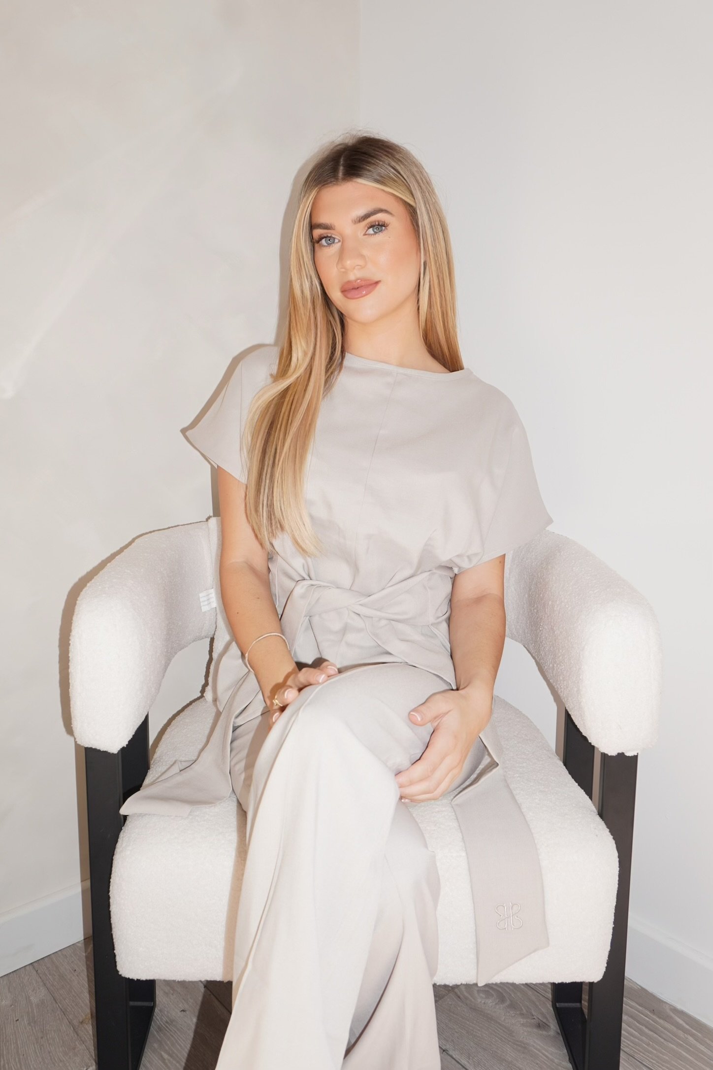 A young woman with long blonde hair and blue eyes sits on a white upholstered chair against a plain wall, wearing a light-colored dress.