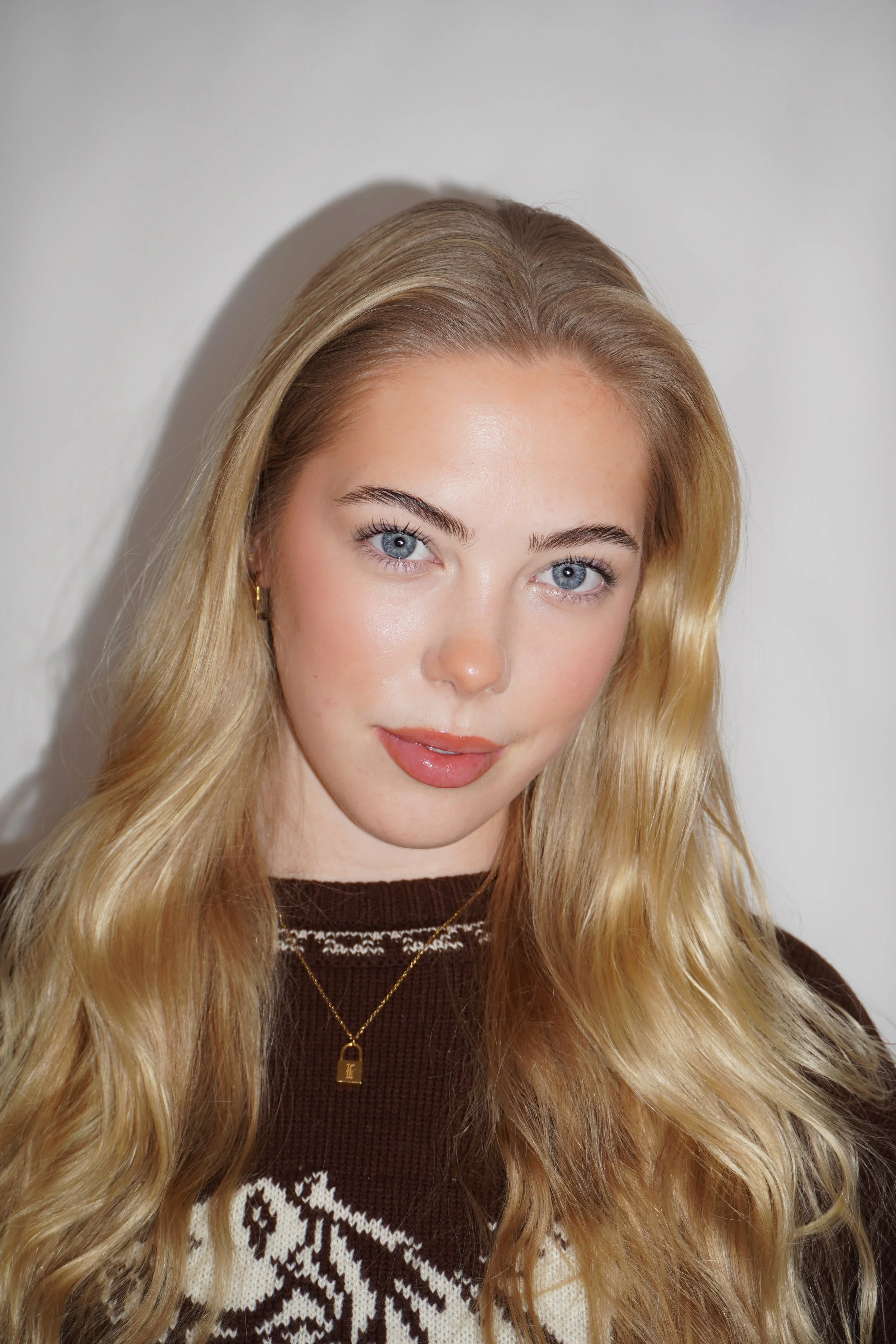 A young woman with long blonde hair, blue eyes, and light skin, wearing a brown and beige patterned sweater and a gold necklace with a small pendant, standing against a plain white wall.