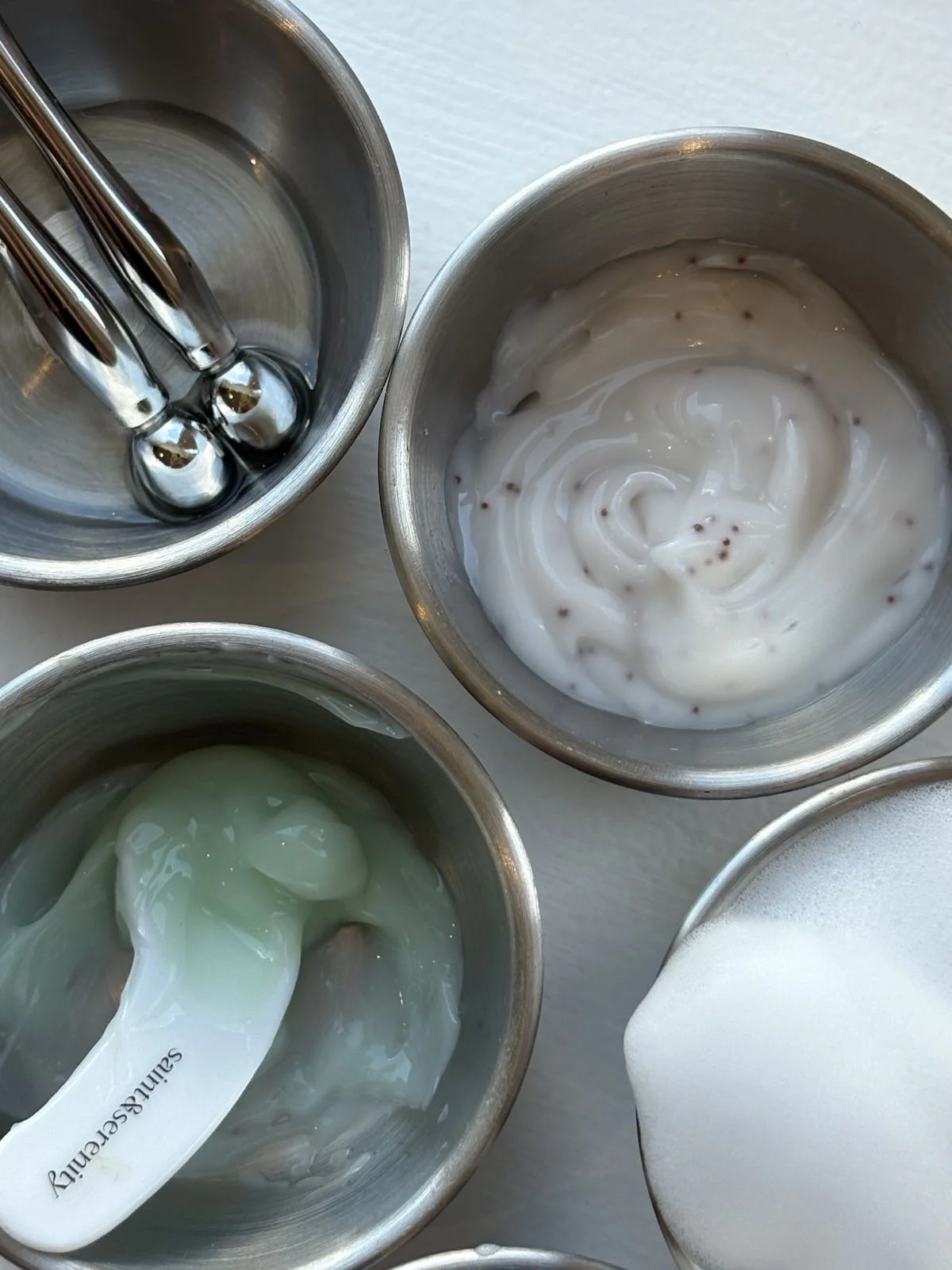 Three small stainless steel bowls with different textures of yogurt or custard, and two identical silver ice cream scoops resting inside the bowls.