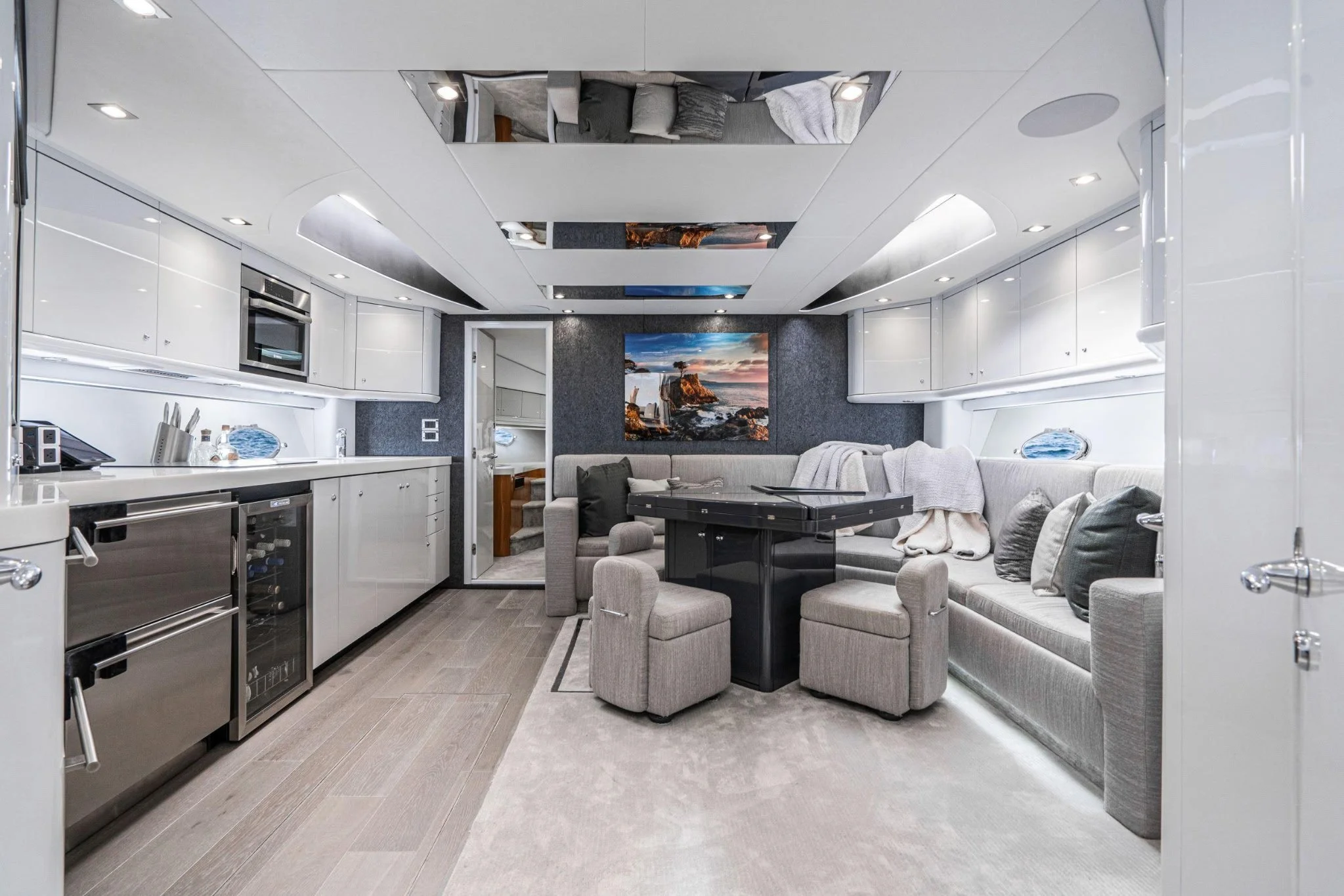 Interior of a modern yacht with a kitchen area on the left, a dining area with a U-shaped sofa and an ottoman in the center, and a ceiling with reflective panels. The walls are white, and there is a painting of a coastal scene on the back wall.