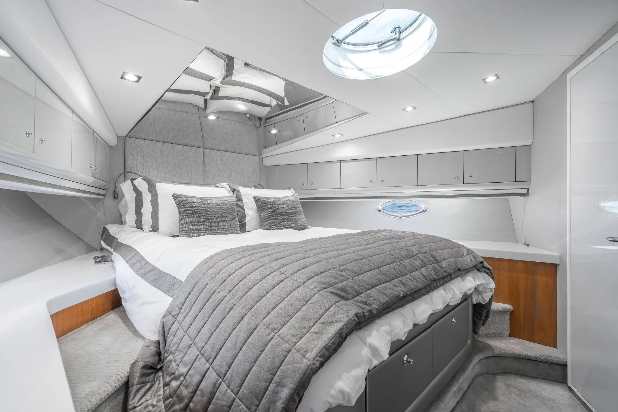 Interior of a modern yacht cabin with a bed, decorative pillows, built-in cabinets, and small windows, in a sleek, gray and white color scheme.
