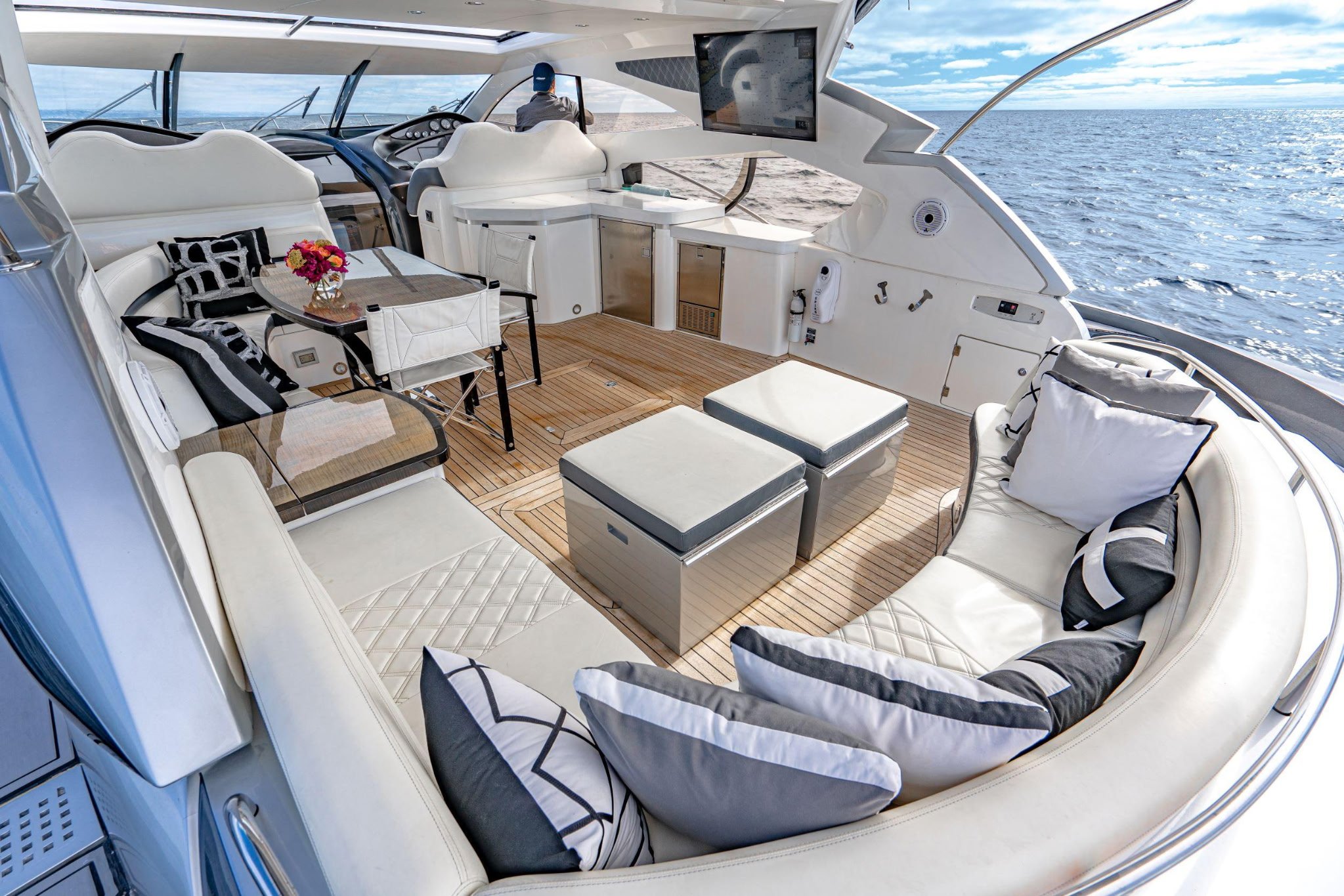 Top deck seating area of a luxury yacht with cushioned white and black pillows, a dining table, and a view of the ocean under a blue sky.