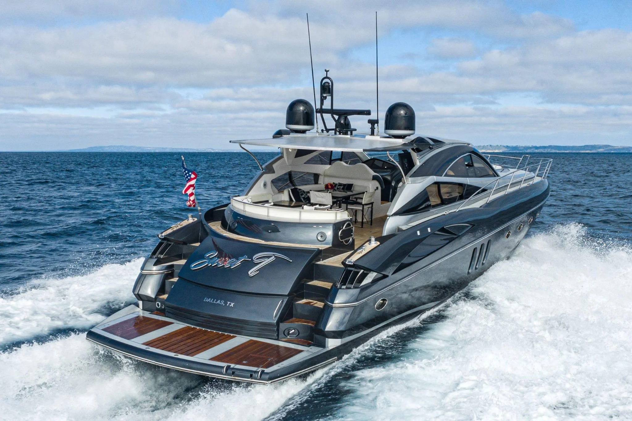 A luxury yacht cruising on open water with a clear sky in the background, featuring a dark hull, modern design, and an American flag at the stern.
