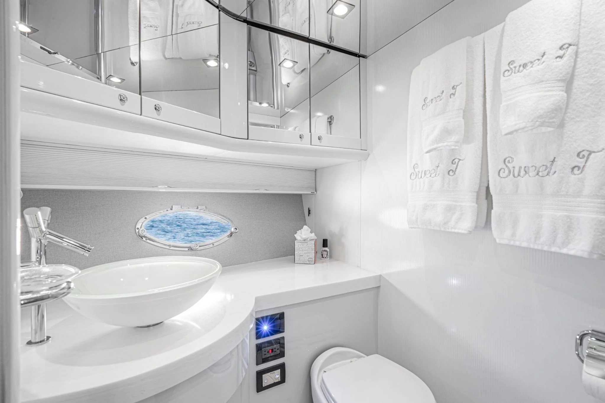 A modern white bathroom with a vessel sink, a small porthole window showing water outside, and two white towels embroidered with 'Sweet T' hanging on the wall.
