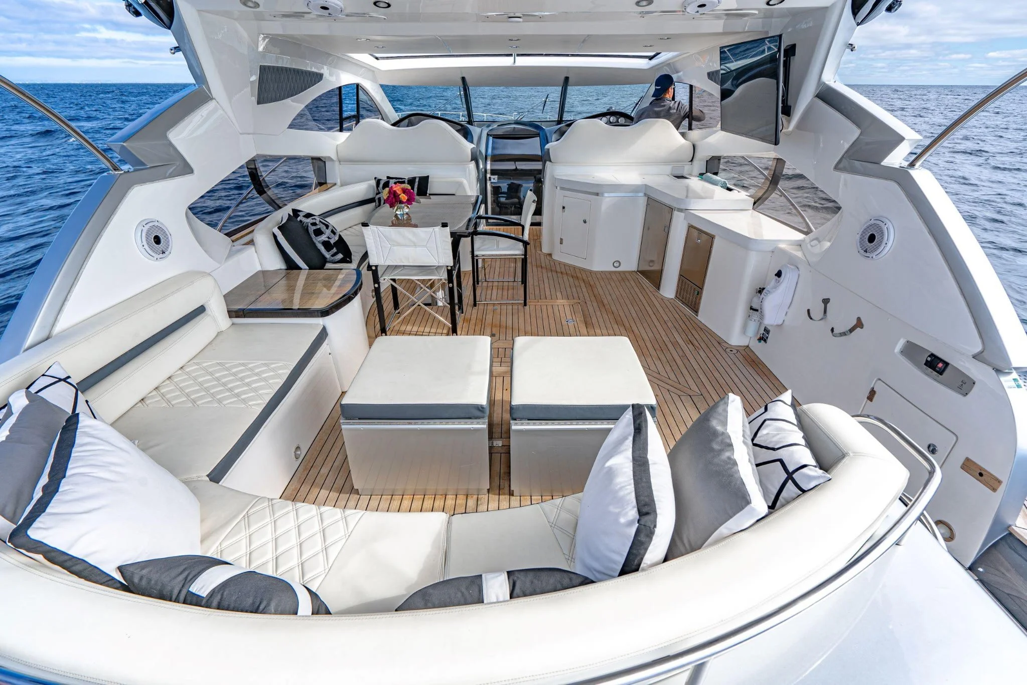 Interior of a yacht with white seating, pillows, a table with a flower arrangement, and a view of the ocean in the background.