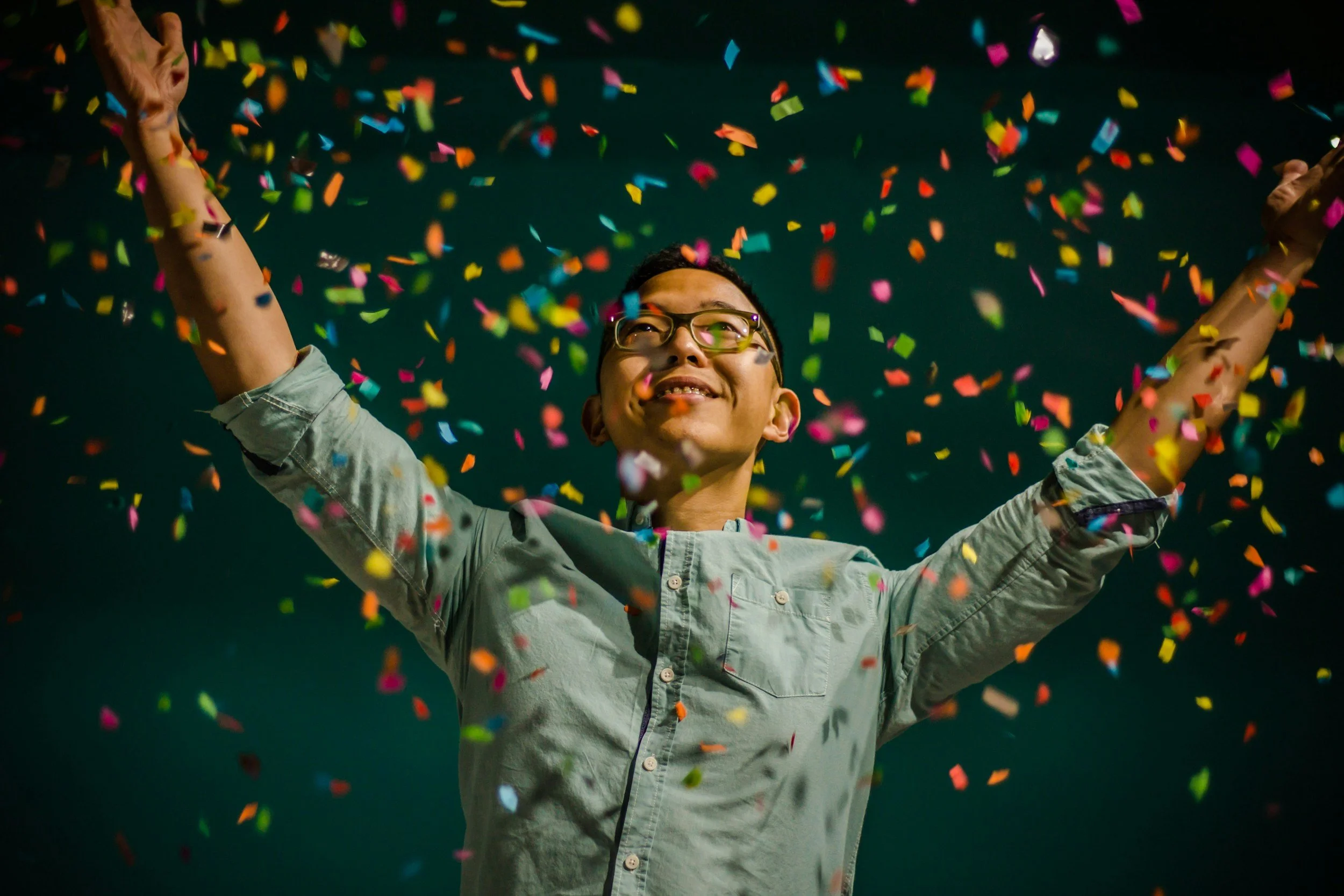 A smiling man wearing glasses and a light green shirt with rolled-up sleeves, celebrating with arms raised amidst falling colorful confetti against a dark background.