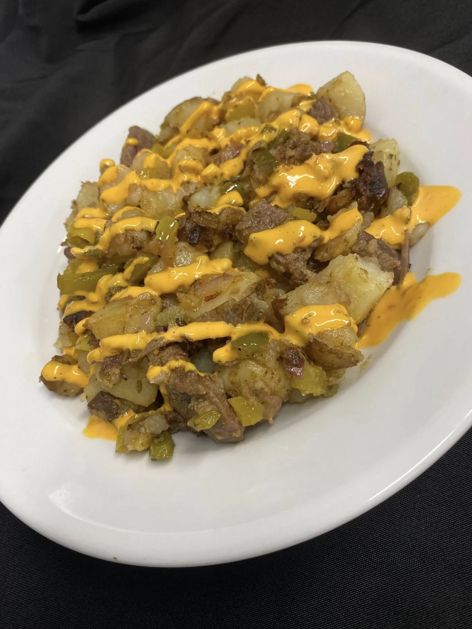 A white oval plate filled with breakfast hash, including diced potatoes, cooked ground meat, green peppers, and cheese, drizzled with melted cheddar cheese.